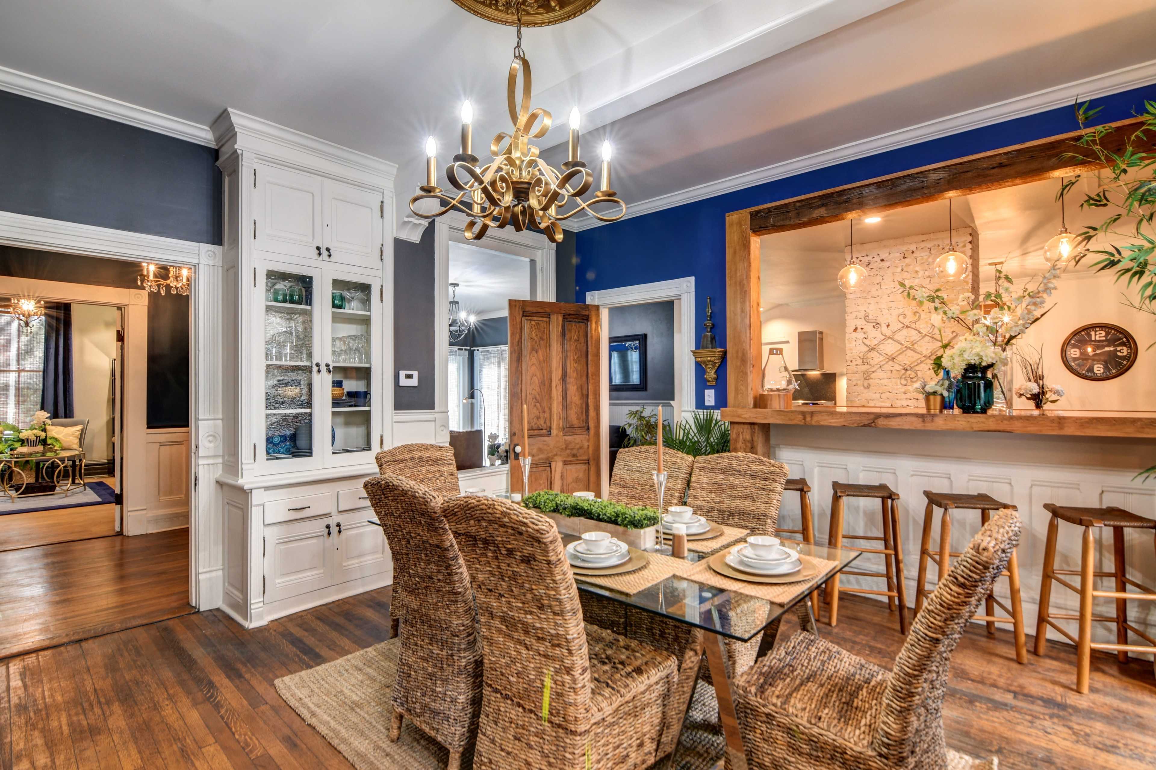 A dining area features a glass table surrounded by wicker chairs, with a bar counter in the background and a chandelier hanging overhead.
