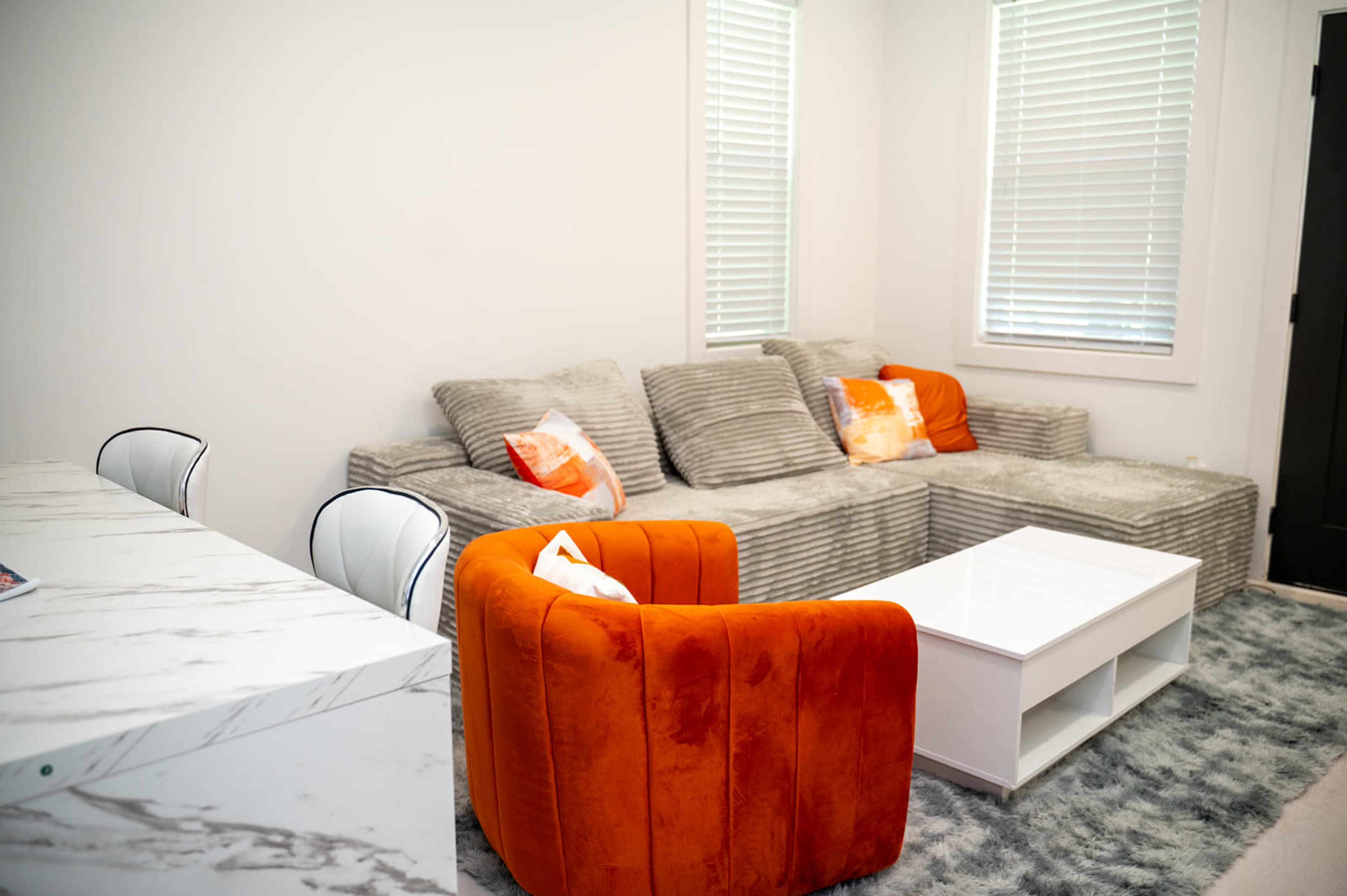 The image shows a modern living room with a striped couch, two orange chairs, a white coffee table, and a marble dining table.