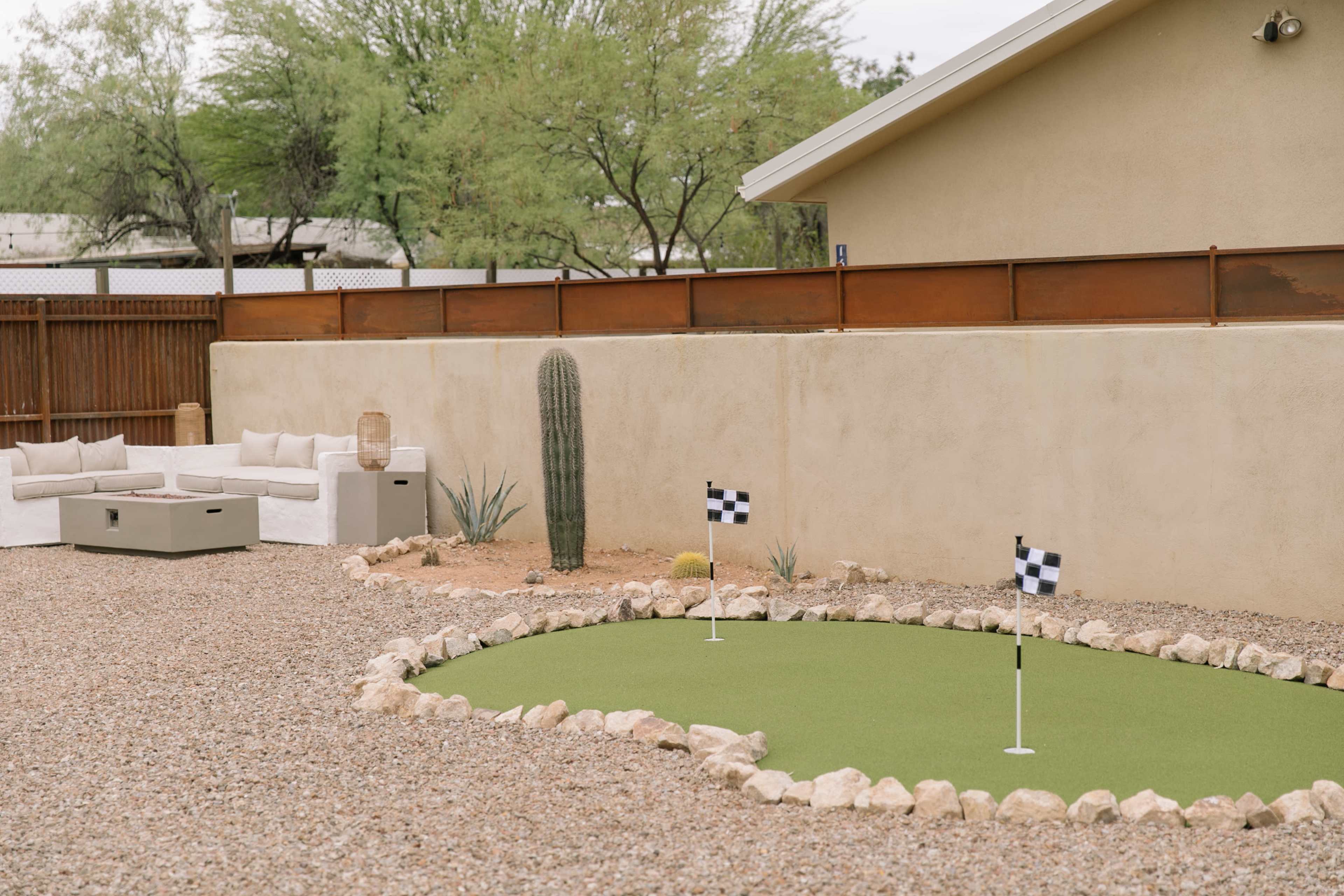 A landscaped backyard featuring a putting green with two flags, surrounded by gravel, cacti, and a seating area.