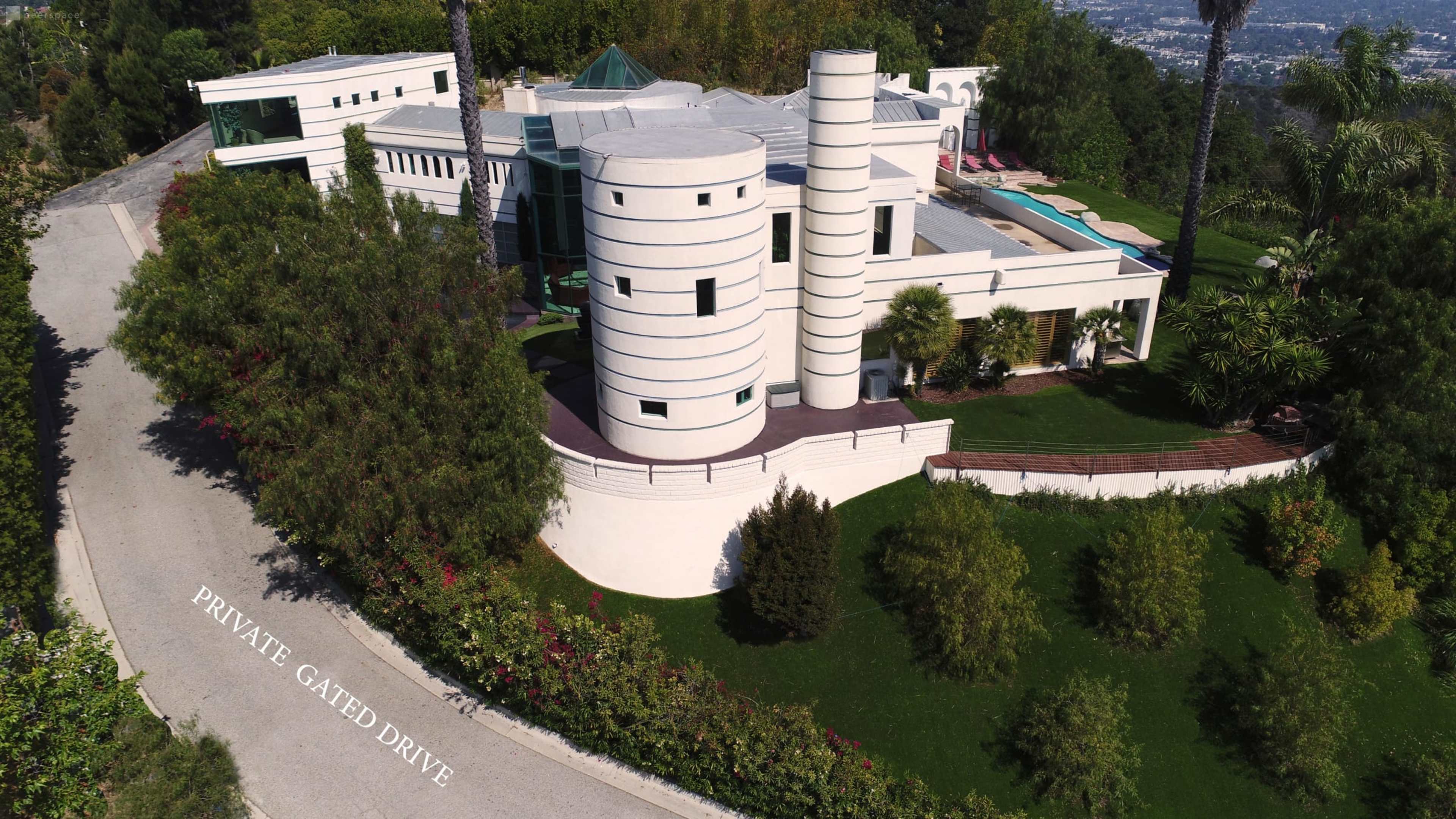 A modern, white cylindrical building with a gated driveway and landscaped greenery surrounding it.