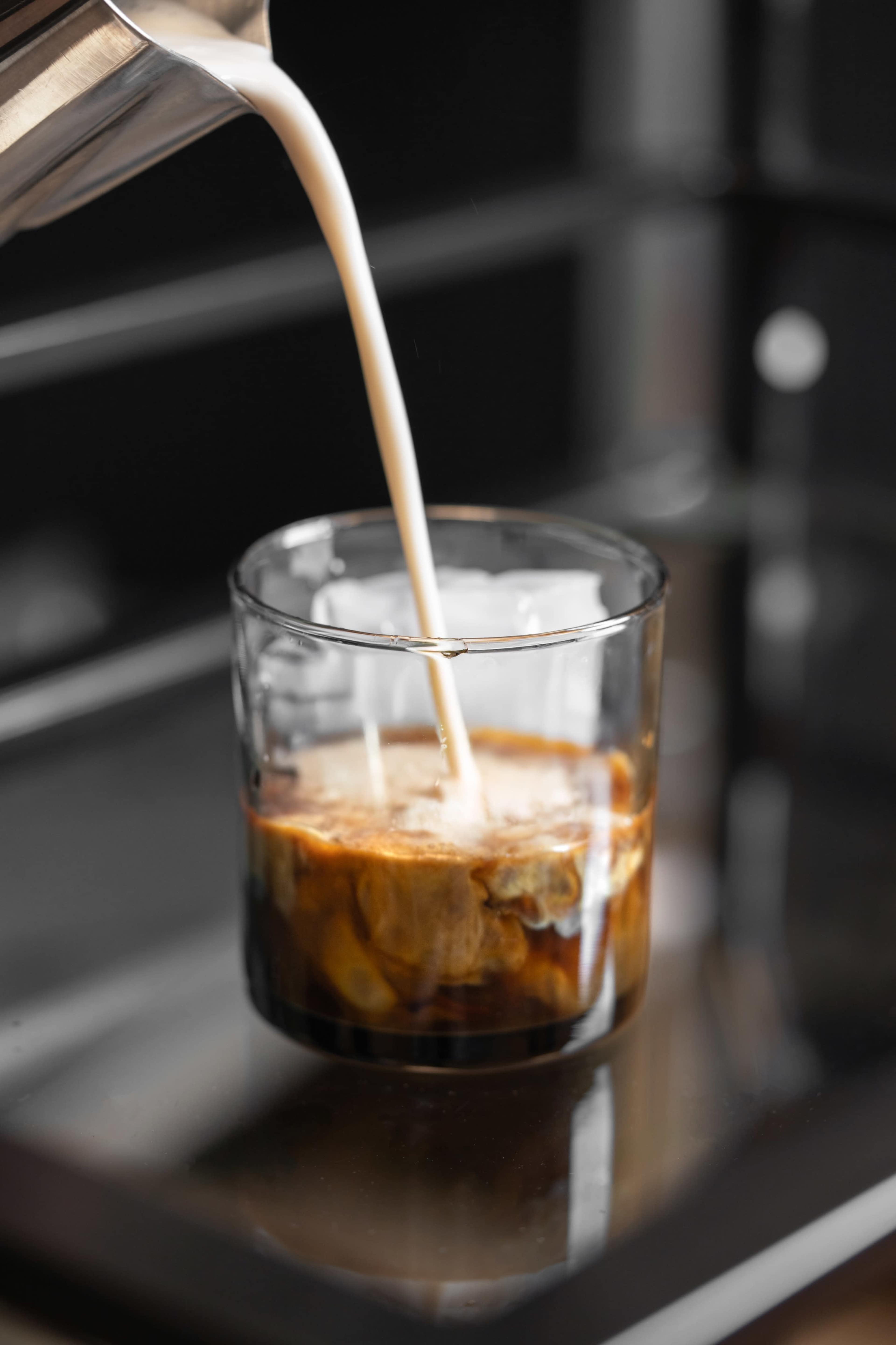 A glass filled with dark liquid receives a pour of white milk from a container above.