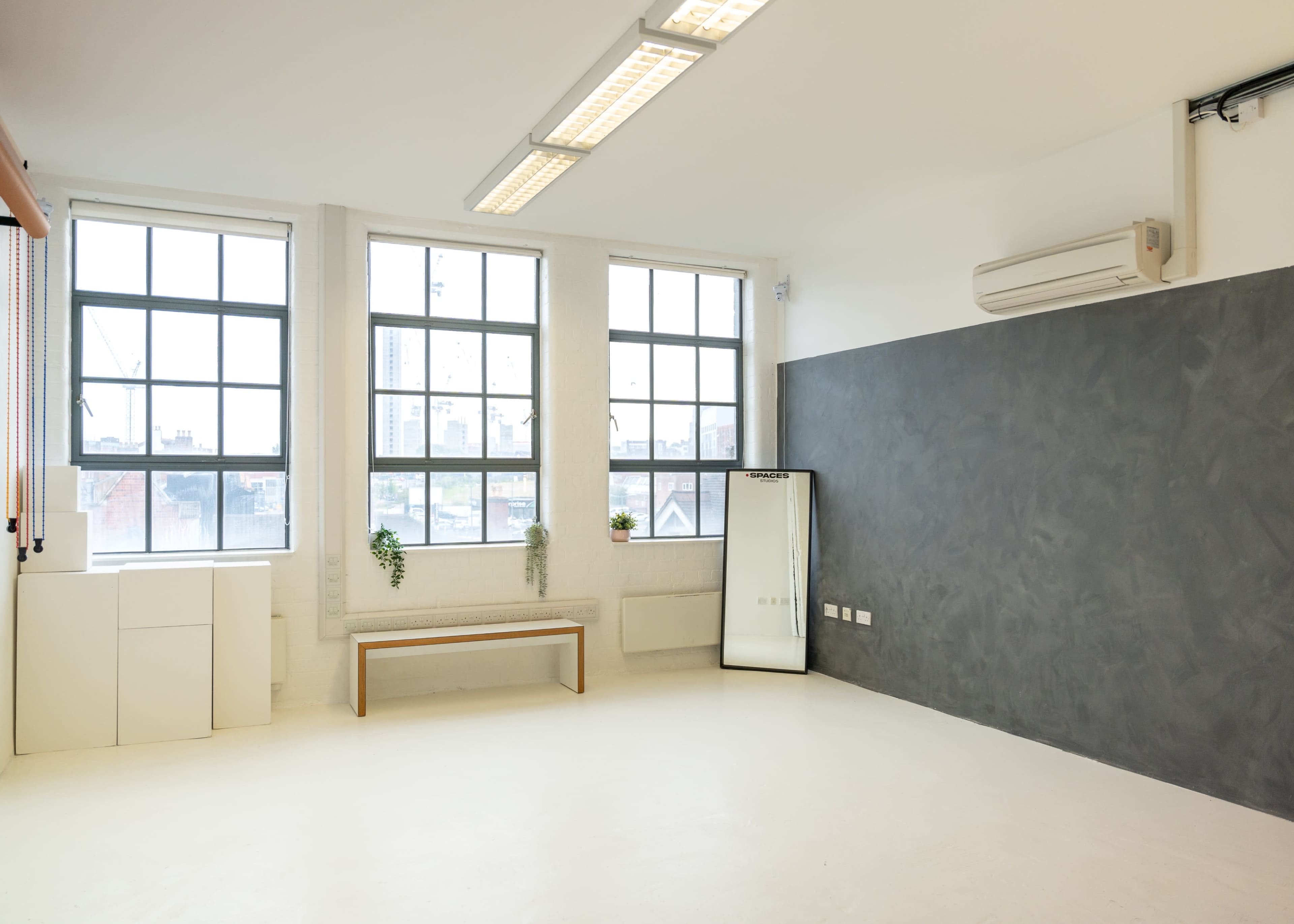 The image shows a bright, minimalistic room with large windows, a bench, a mirror, and a dark accent wall.