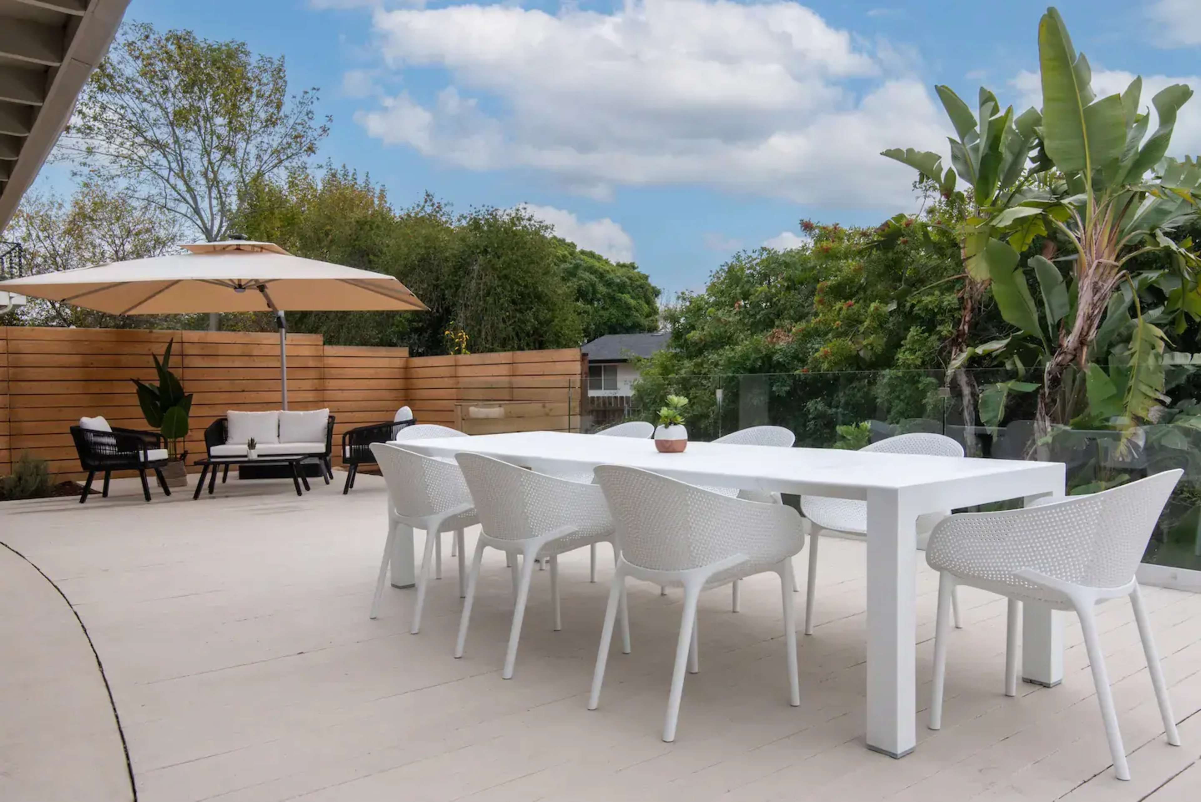A modern outdoor dining area with a white table and eight white chairs, surrounded by green plants and a large umbrella.