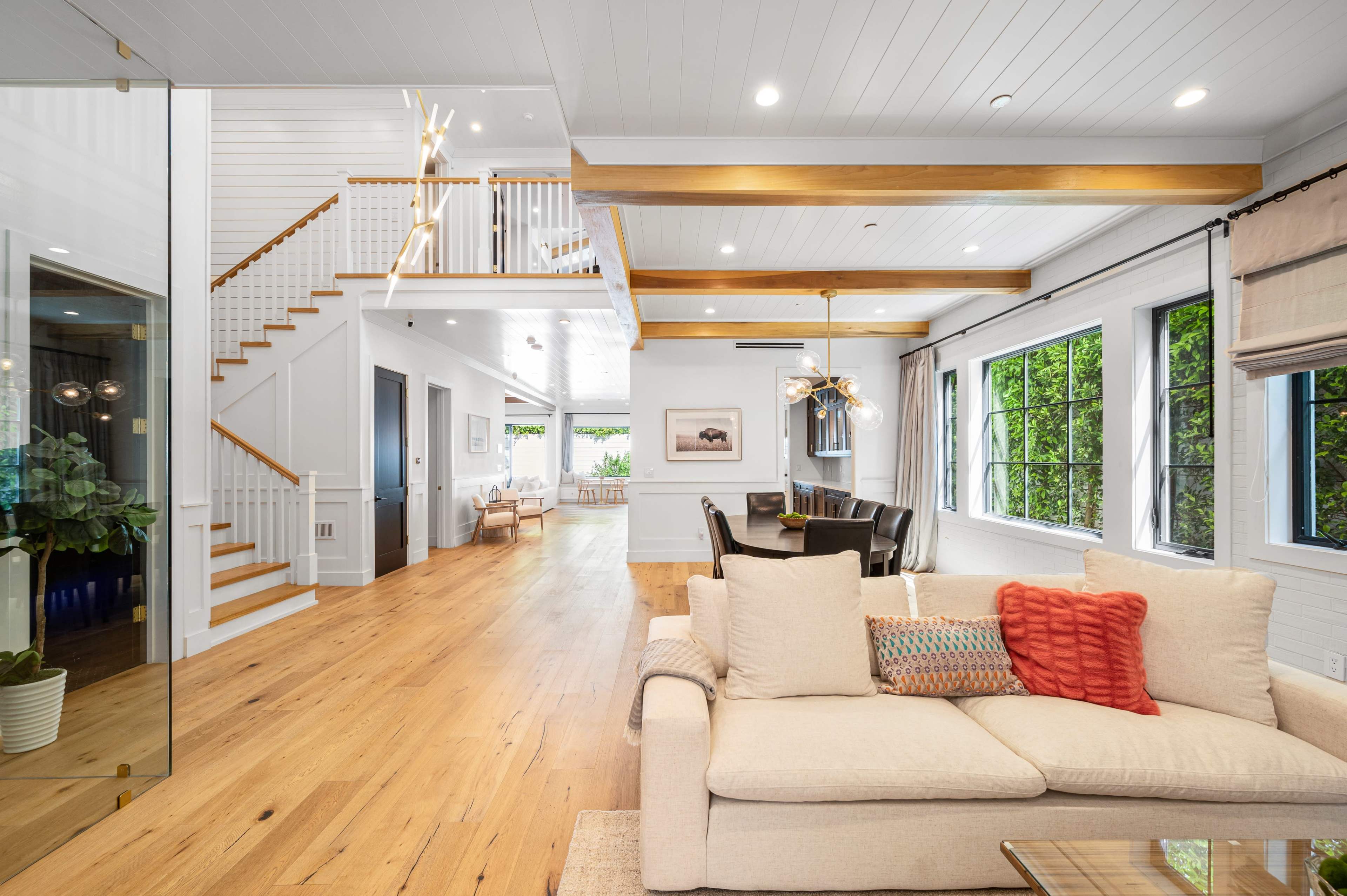 A spacious interior with a living area featuring a beige sofa, a staircase, and large windows that open to a green outdoor space.