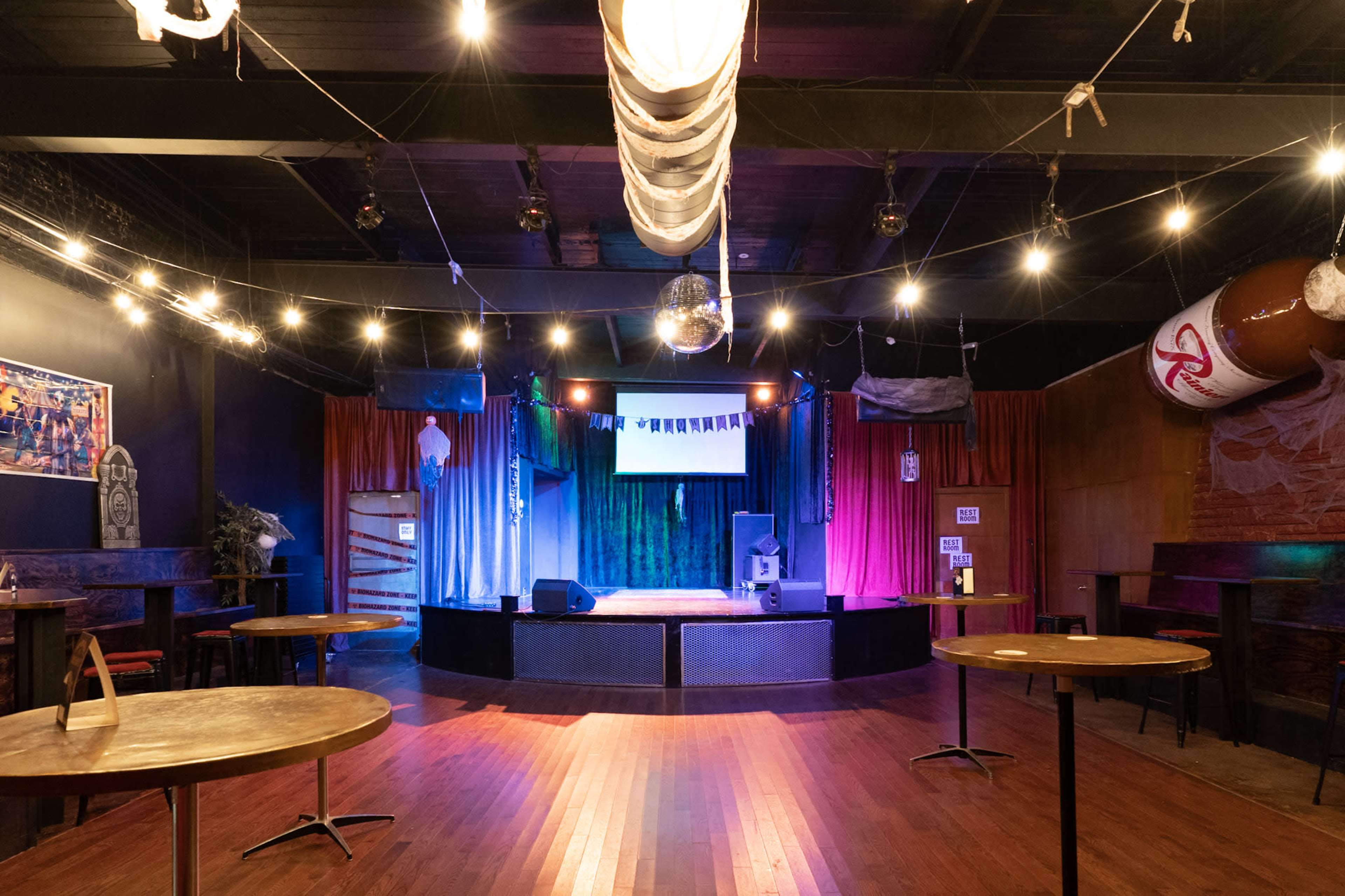 The image shows a dimly lit stage area in a bar or performance venue with empty round tables, colorful curtains, and a screen set up for presentations or performances.
