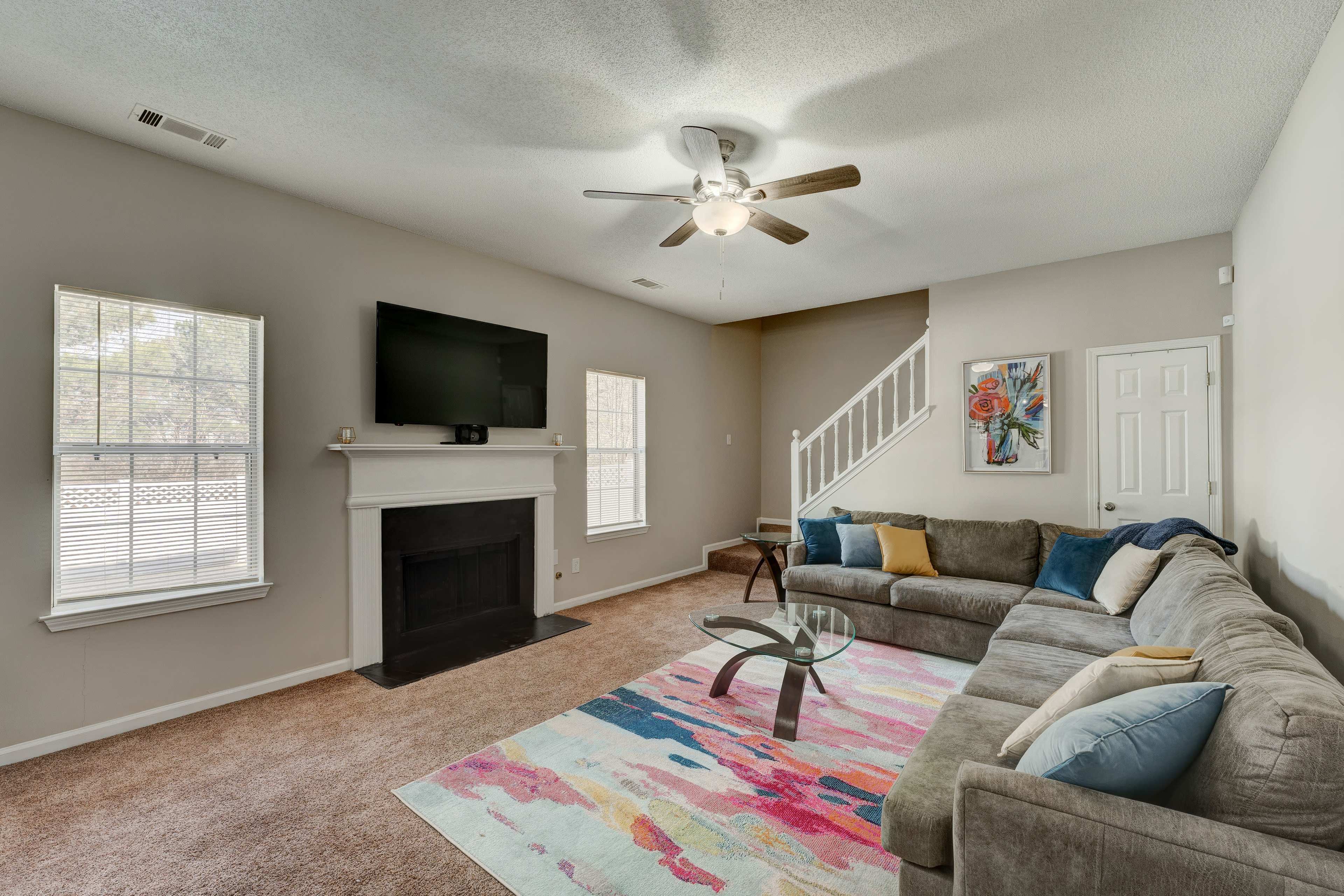 A living room with a sectional sofa, a coffee table, a wall-mounted television, and a staircase, featuring light-colored walls and carpet.