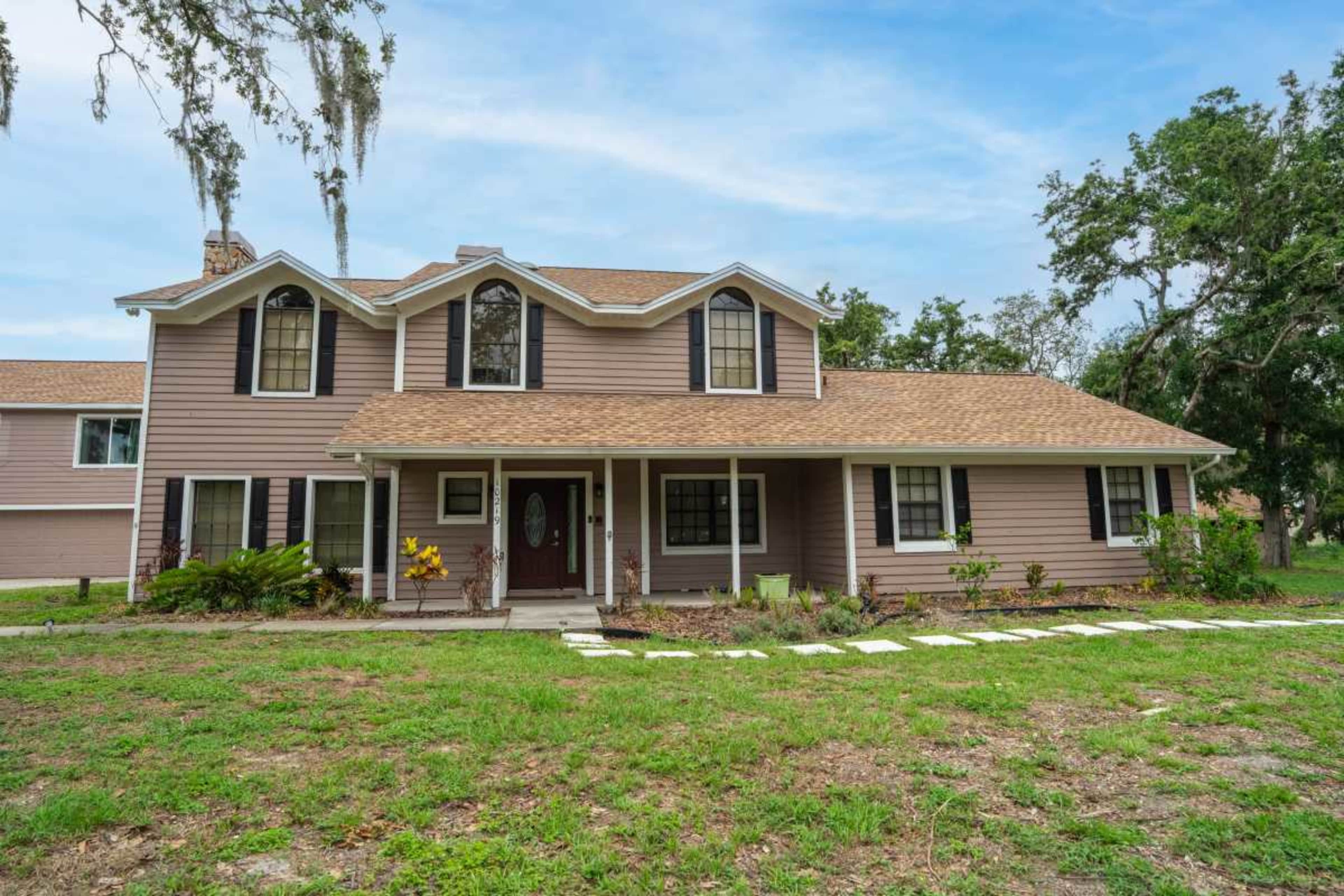 The image shows a two-story house with a brown exterior, a gabled roof, and a front porch, surrounded by a grassy yard.