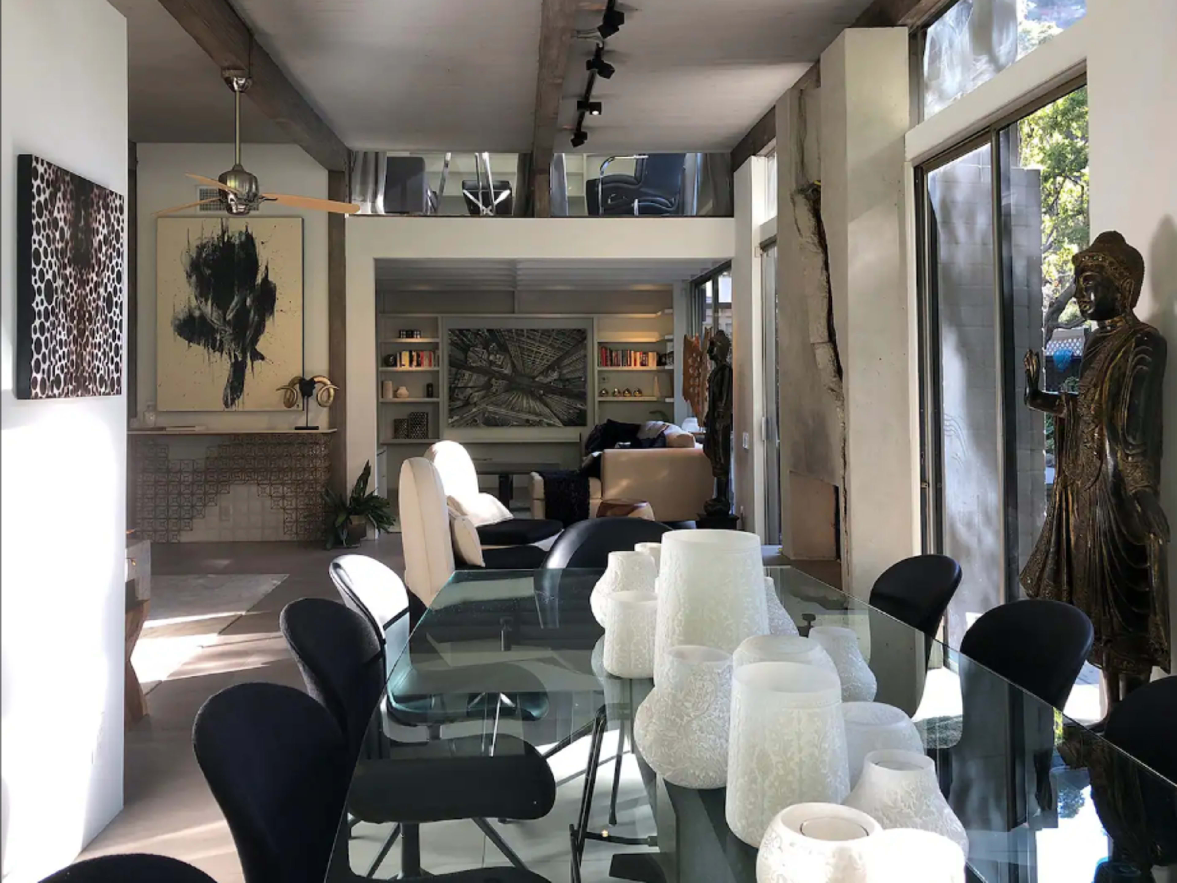 A modern living space features a glass dining table, artwork on the walls, and a mix of furnishings and decorative items throughout.