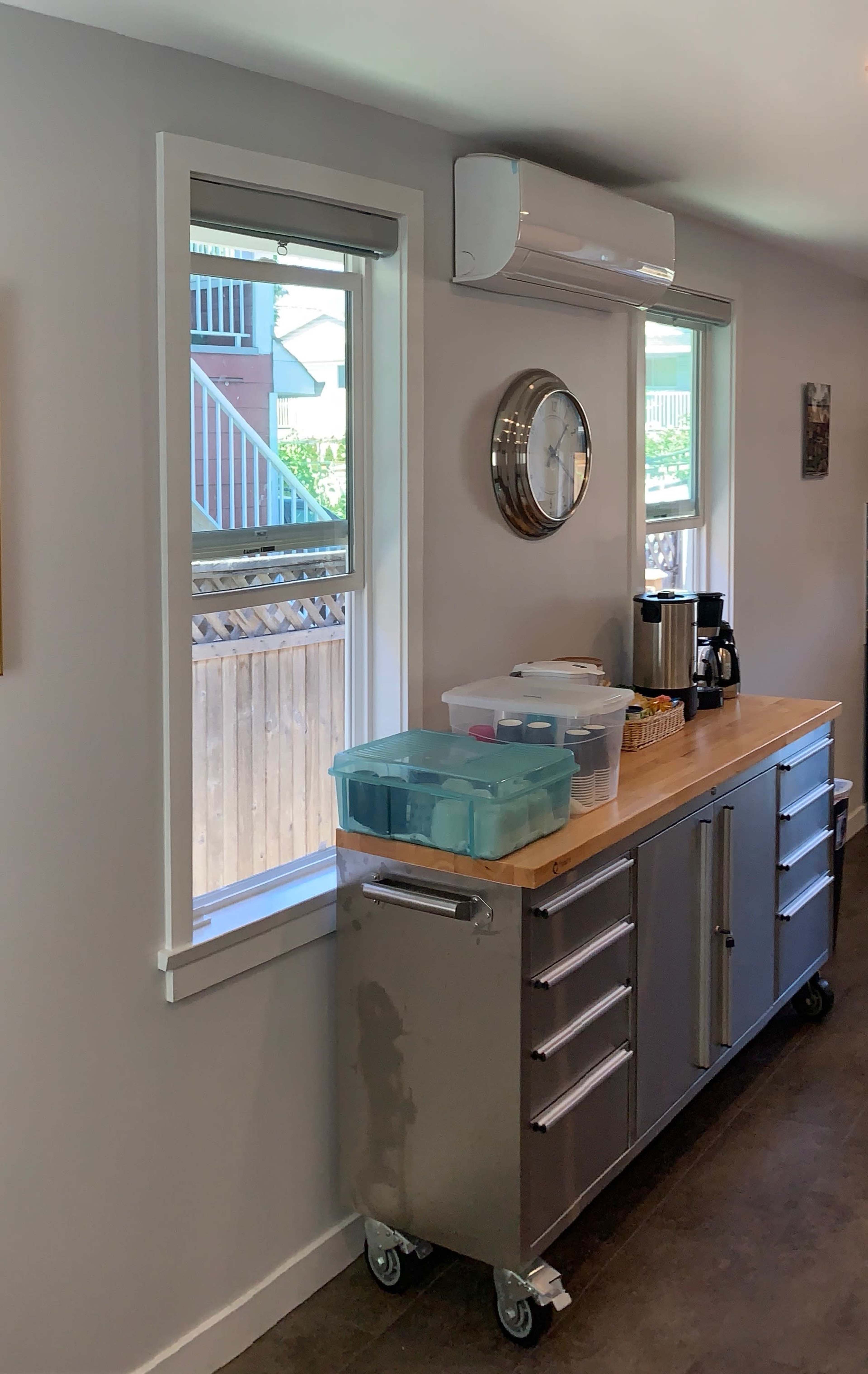 A modern kitchen with a stainless steel cart, a coffee maker, a clock on the wall, and windows allowing natural light to enter.