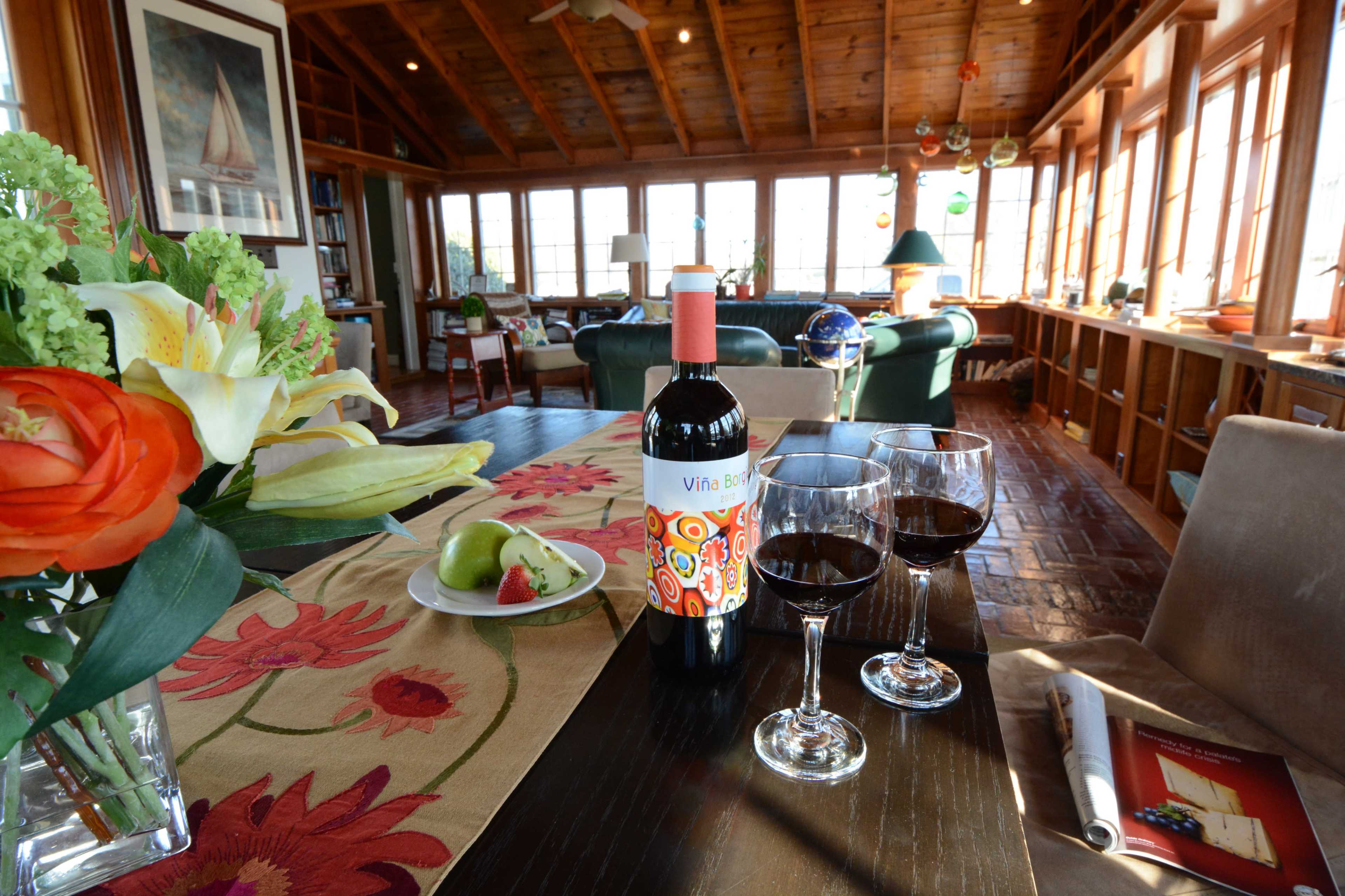 A table is set with a bottle of wine and two glasses, surrounded by flowers and fruit, while a cozy living area with large windows is visible in the background.