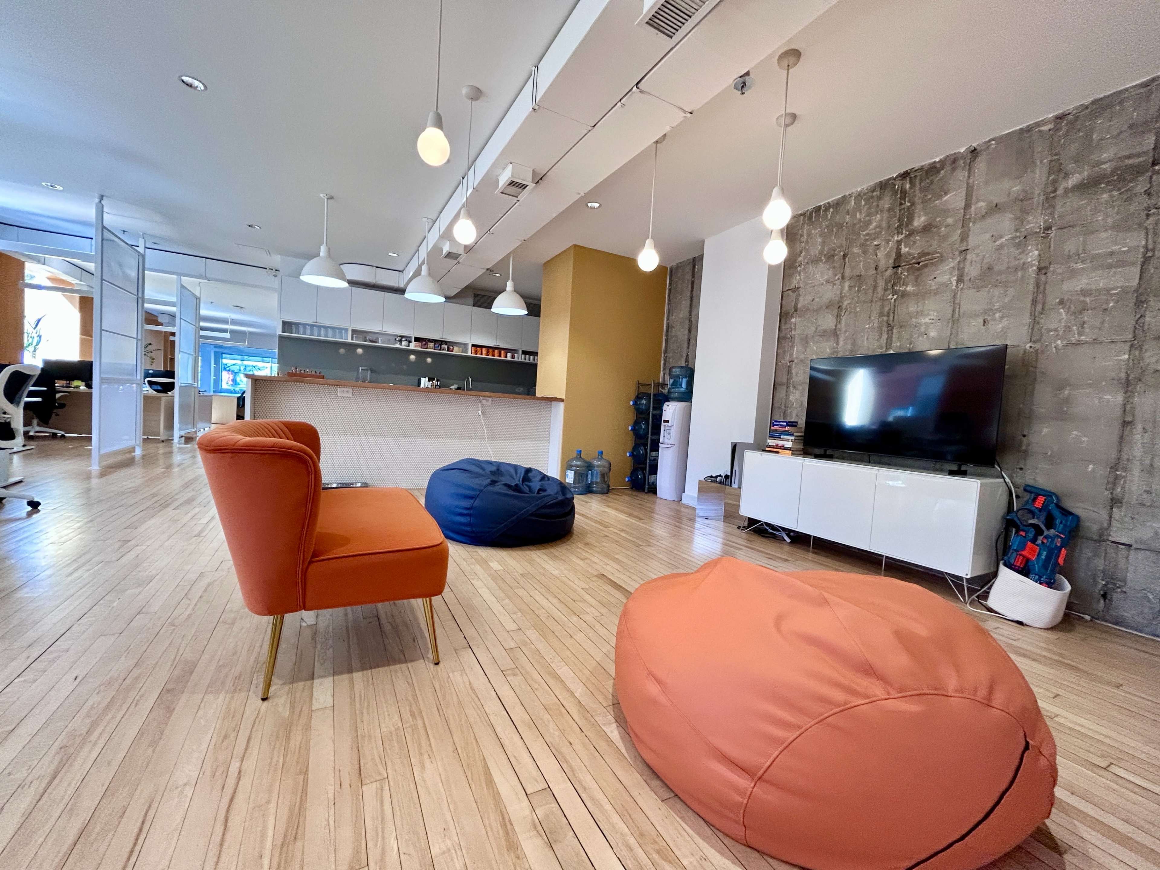 The image shows a modern office space featuring wooden flooring, colorful bean bags, a TV on a white cabinet, and a reception area with pendant lights.