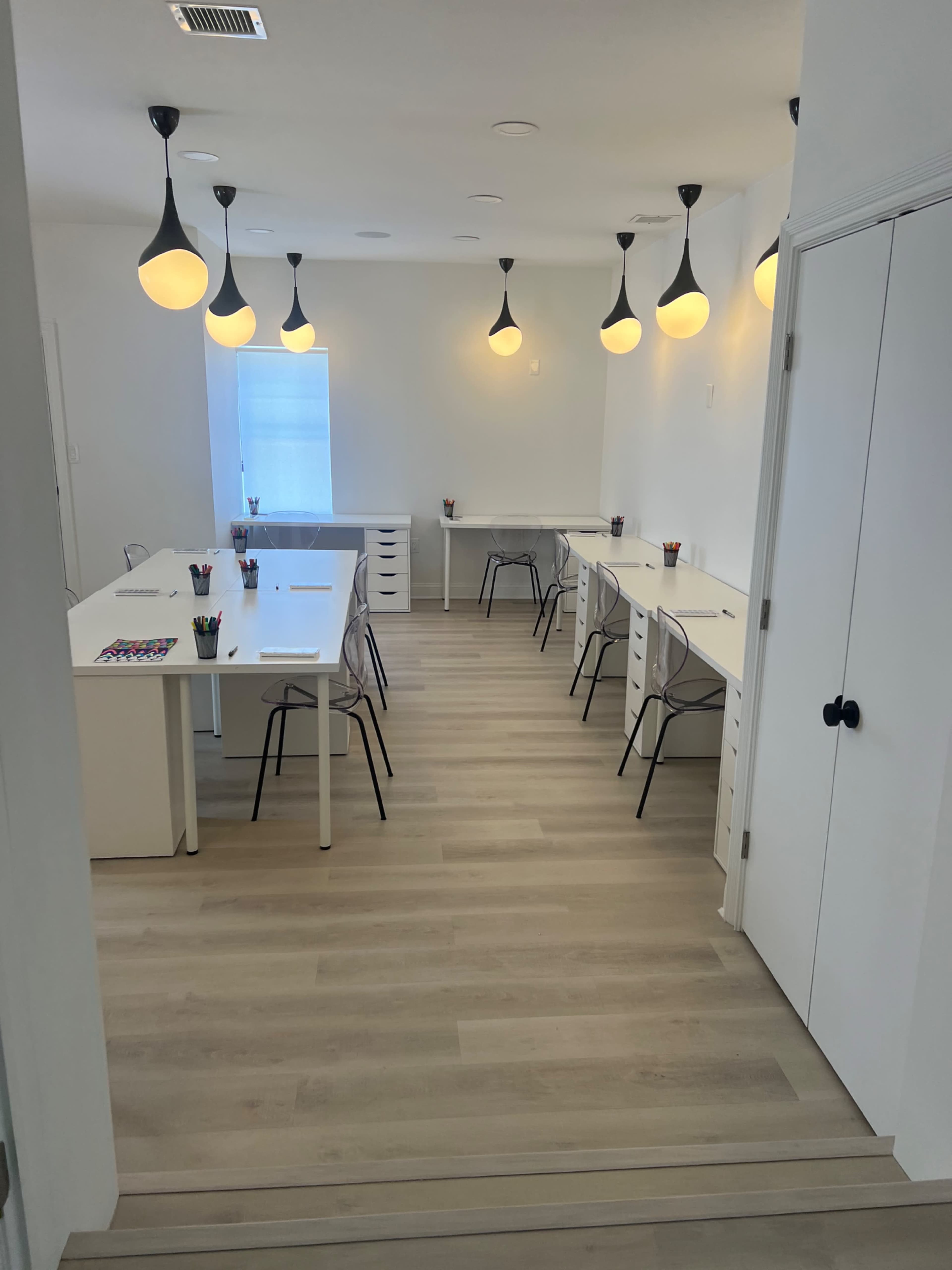 A bright, minimalist workspace with several desks arranged in a spacious room, illuminated by pendant lights.
