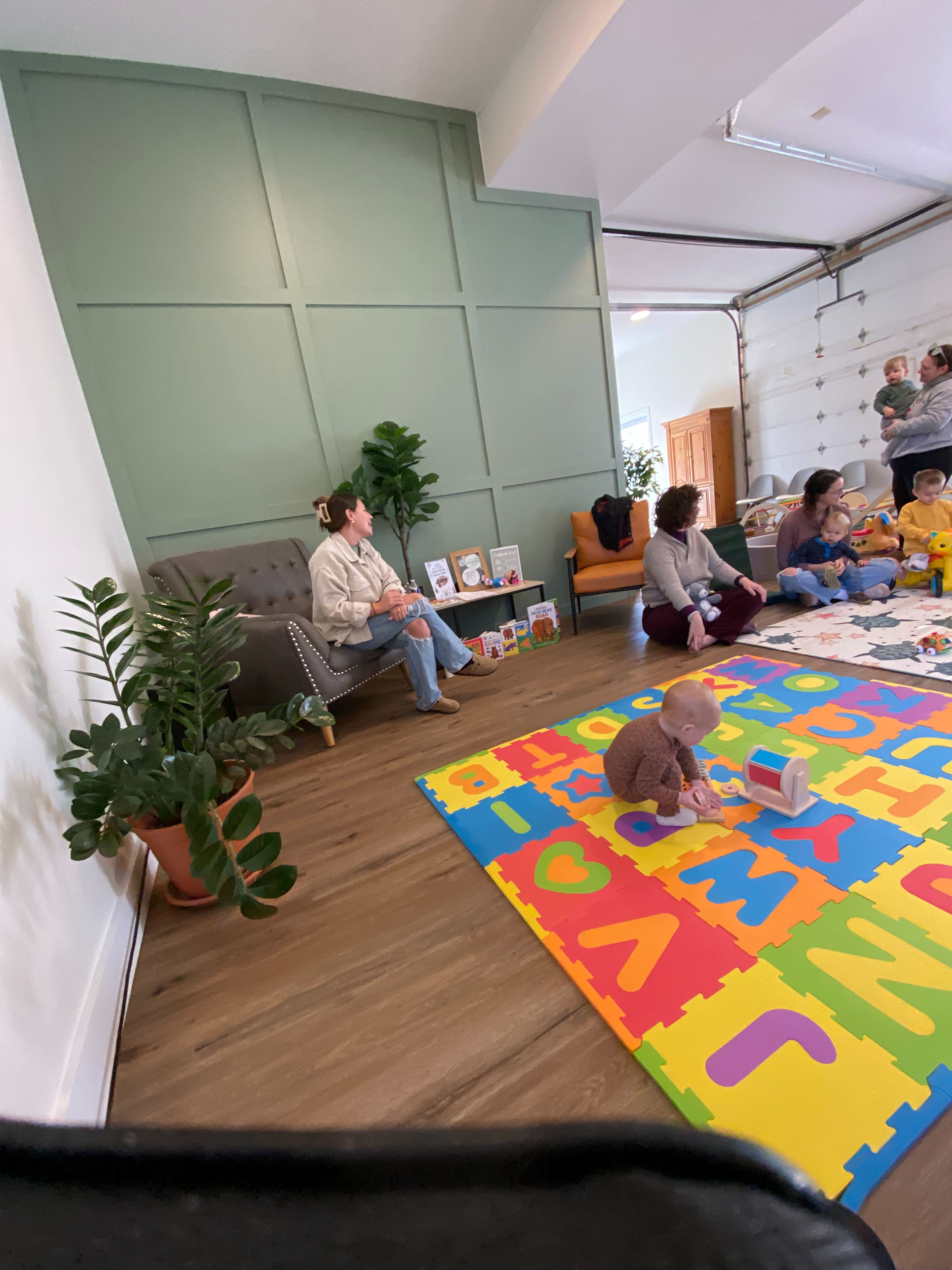 A spacious room features a colorful alphabet play mat, a seated woman beside a plant, and a group of people engaged in activities on the floor.