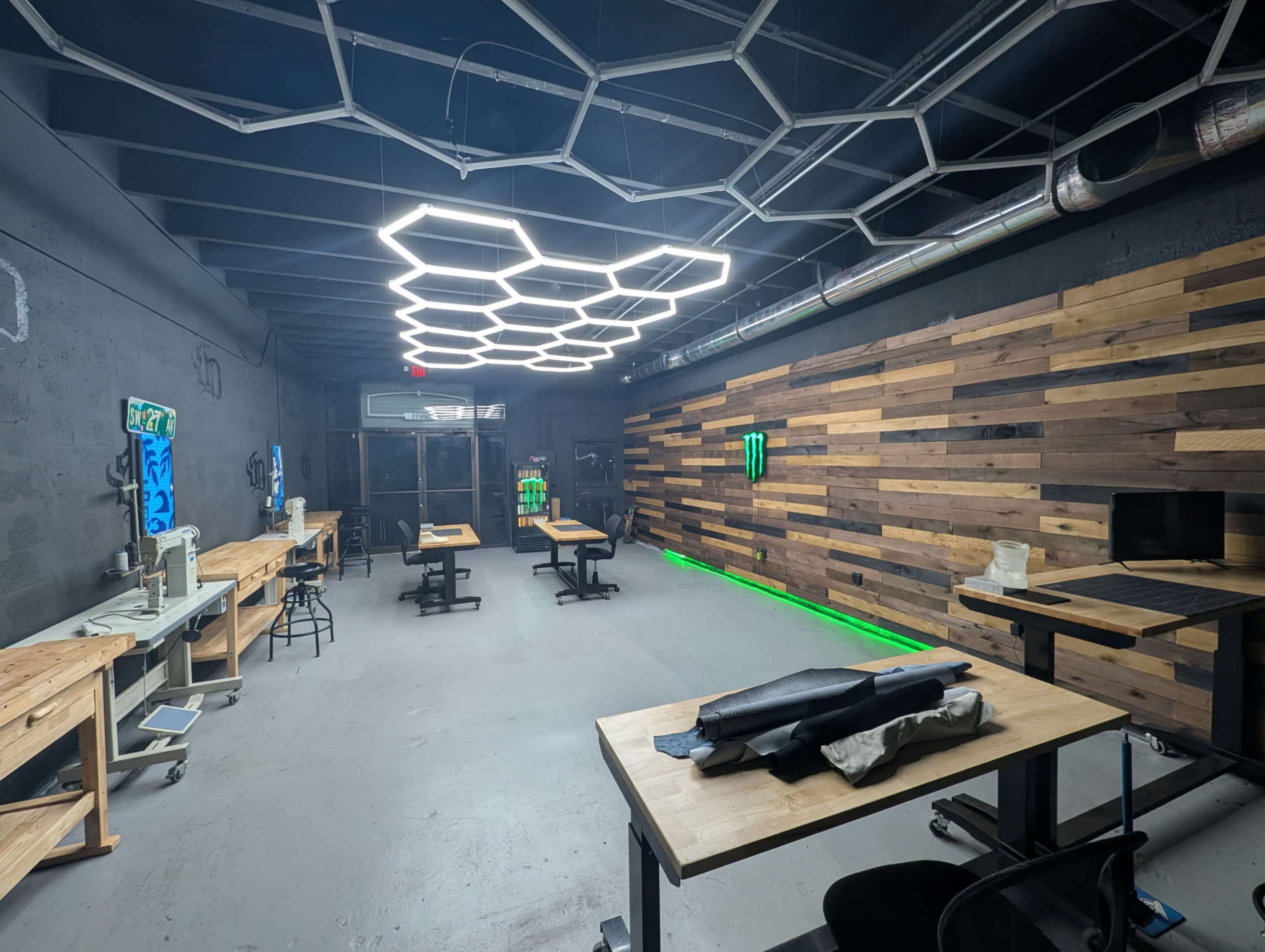 The image shows an industrial-style workspace featuring light wooden tables, a wooden wall, and modern hexagonal ceiling lights, with some green lighting in the background.