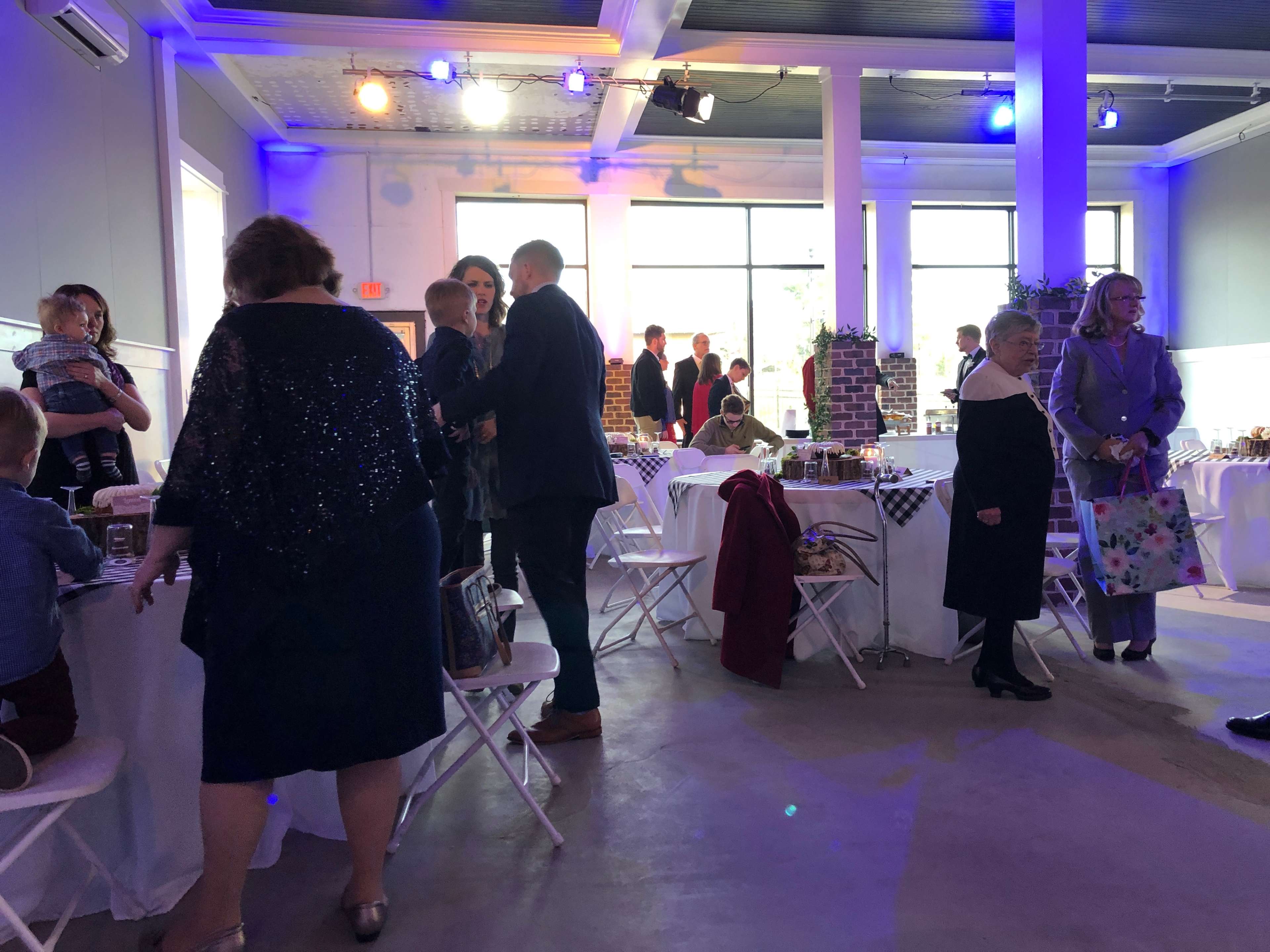A spacious event venue with guests interacting and seated at white tables, while a few people stand around, surrounded by decorations and a large window letting in natural light.
