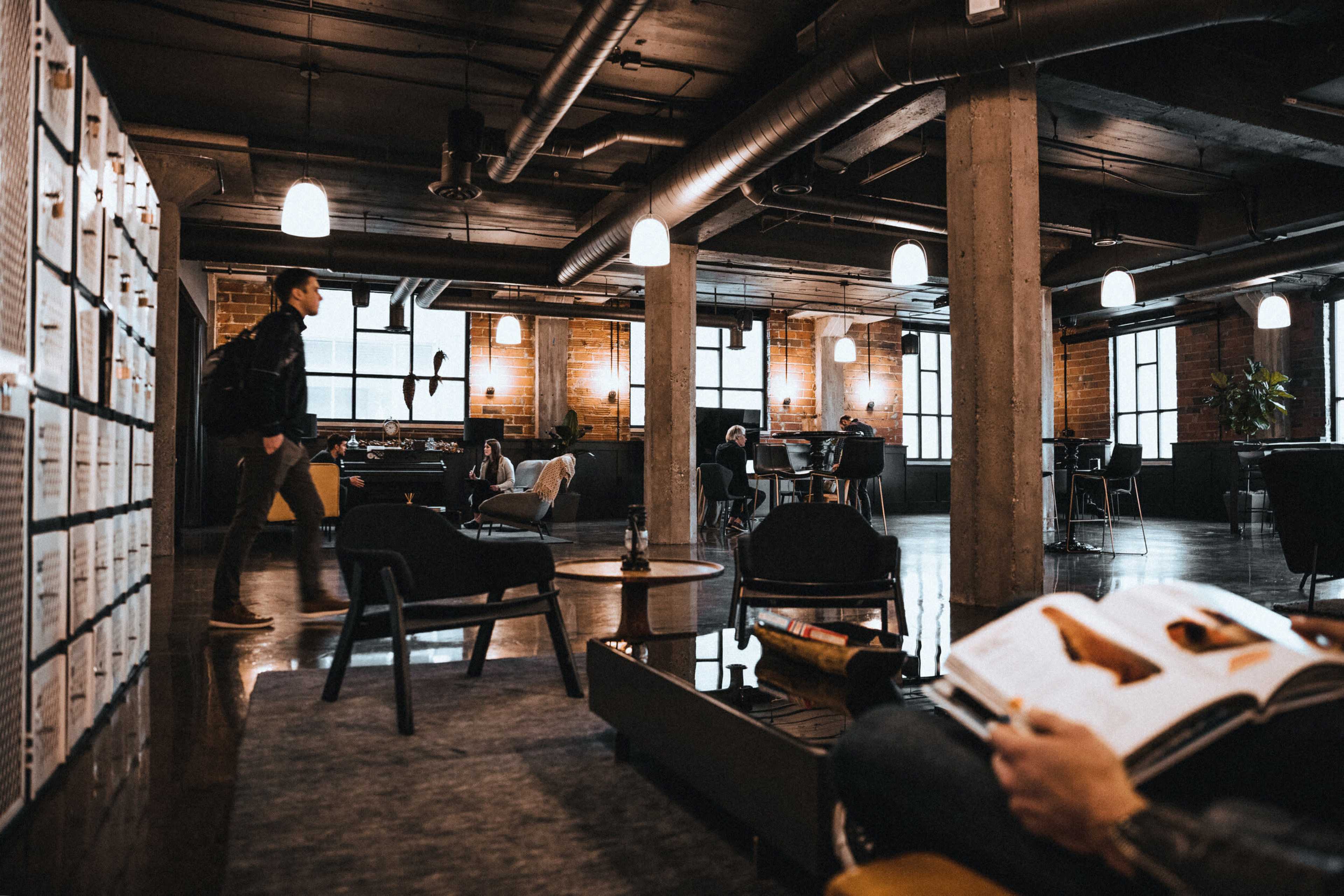 A modern co-working space features a mix of seating areas, large windows, and exposed brick walls, with people engaged in various activities.