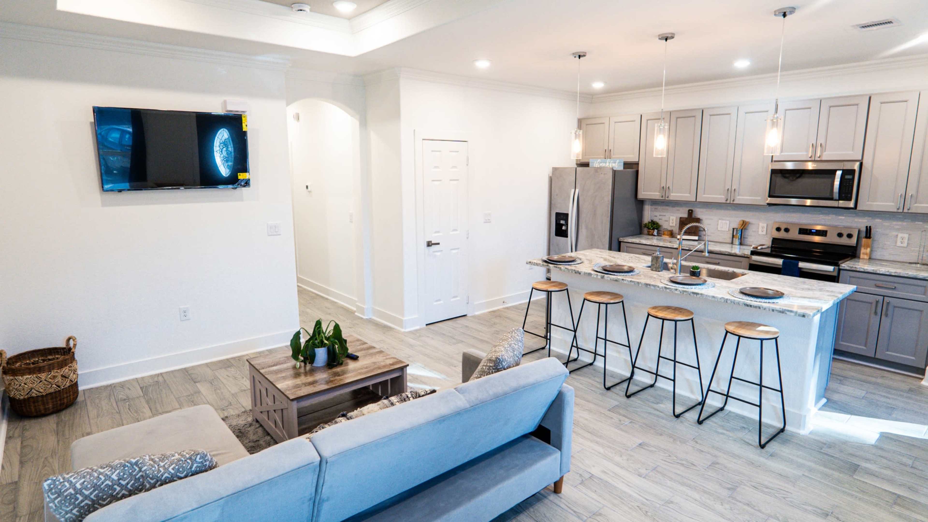 The image shows a modern open-concept living area with a kitchen, featuring a sofa, a coffee table, and barstools at the kitchen island.
