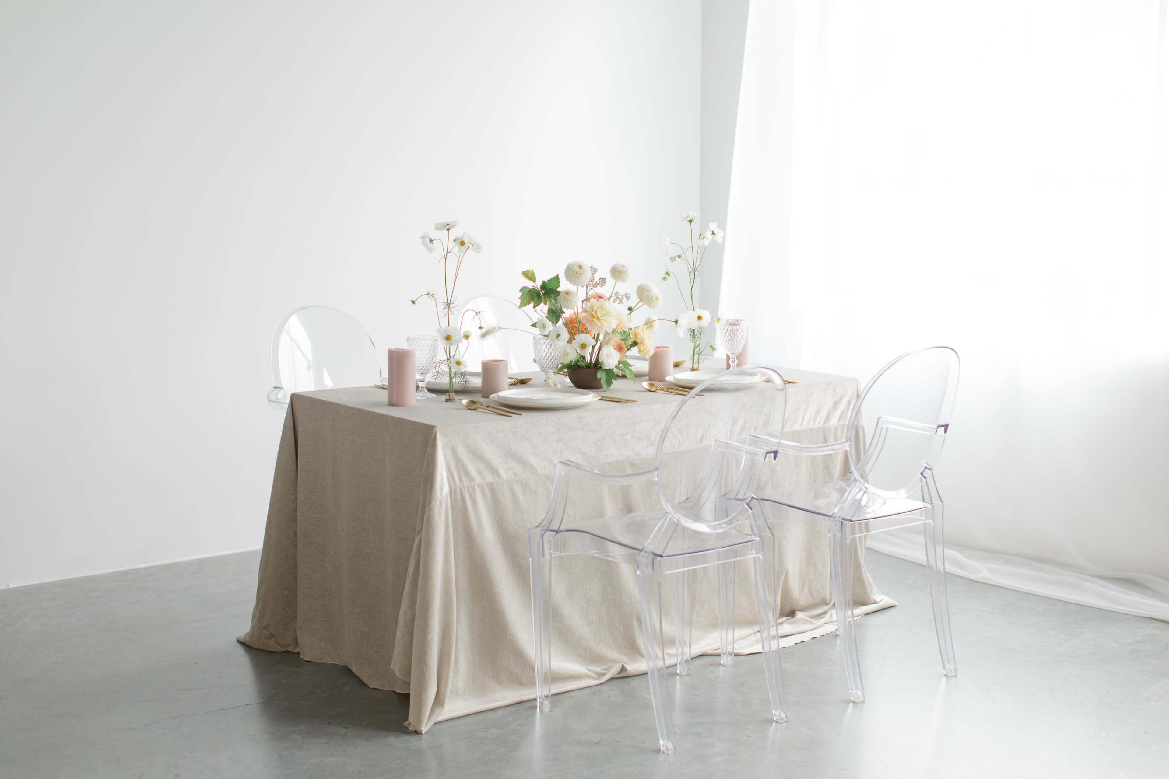 A dining table set with a neutral tablecloth, plates, and vases of flowers, surrounded by transparent chairs.