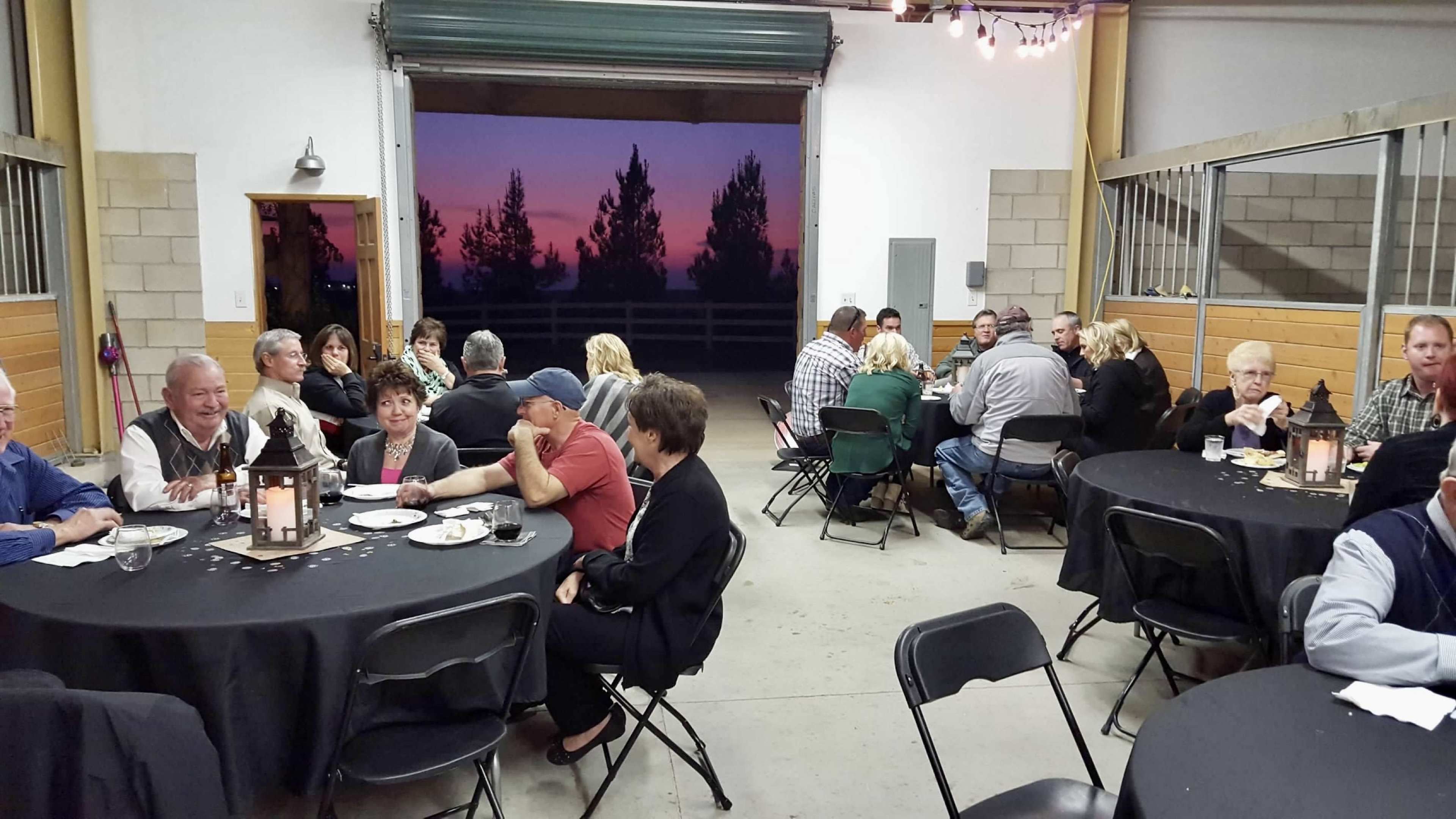 A group of people is seated at tables during an indoor gathering, with a sunset visible through an open garage door in the background.