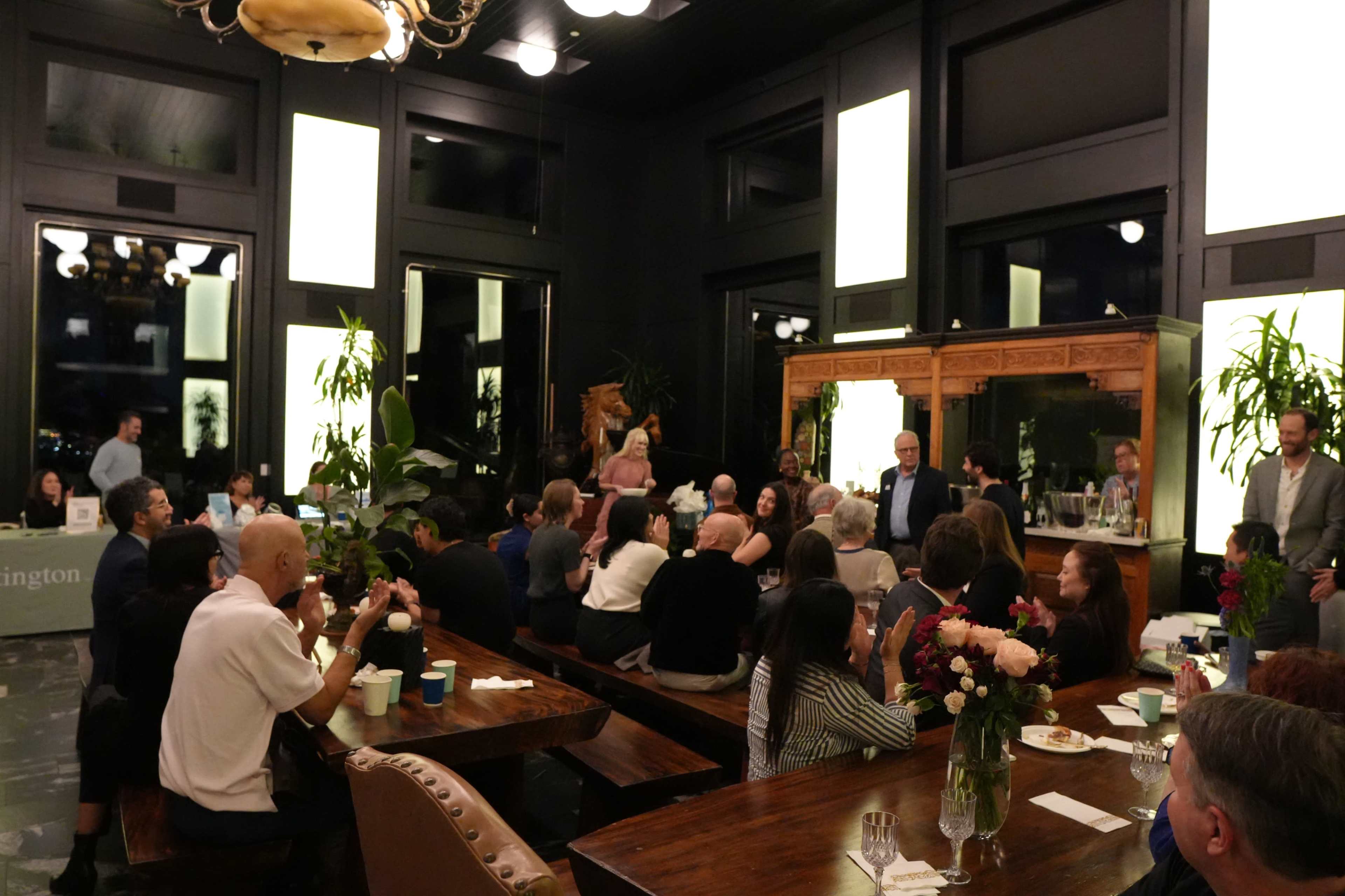 The image shows a large indoor gathering with people seated at long wooden tables, socializing in a well-lit room decorated with plants and large windows.
