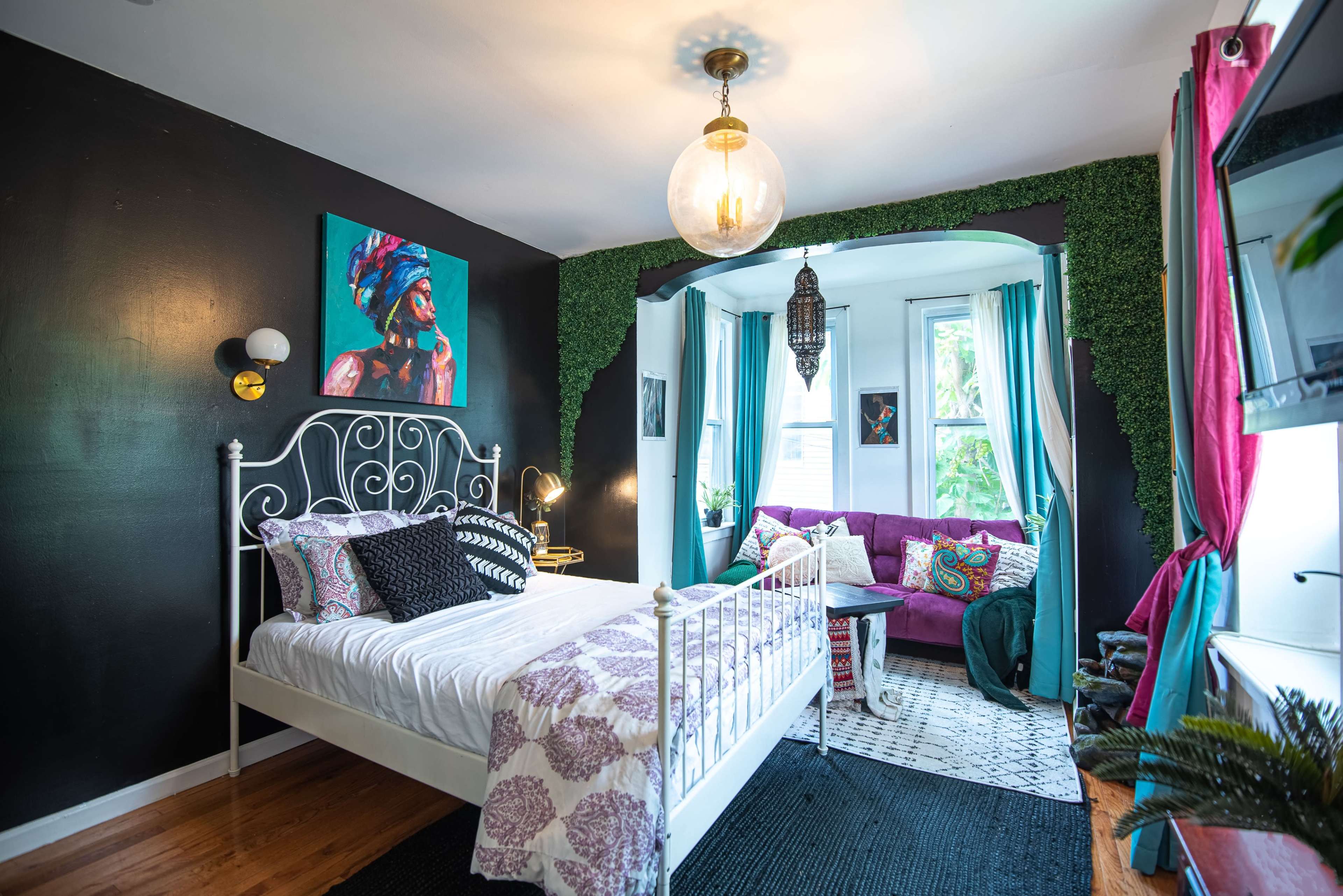 The image shows a stylish bedroom featuring a white metal bed, dark walls, vibrant artwork, and a cozy seating area with plants and decorative textiles.
