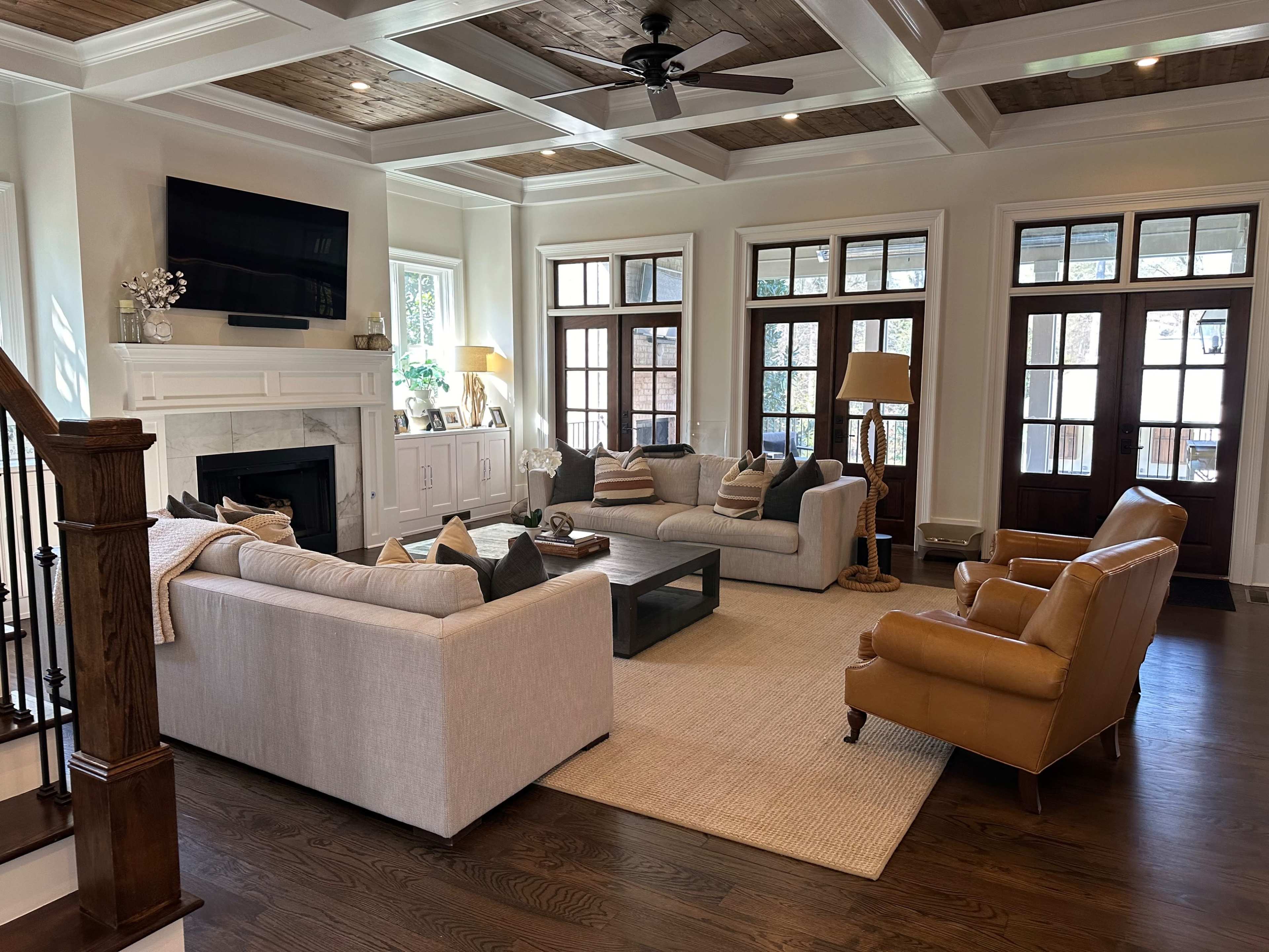 The image shows a spacious, well-lit living room with two light-colored sofas, a coffee table, and large windows, framed by a coffered ceiling.