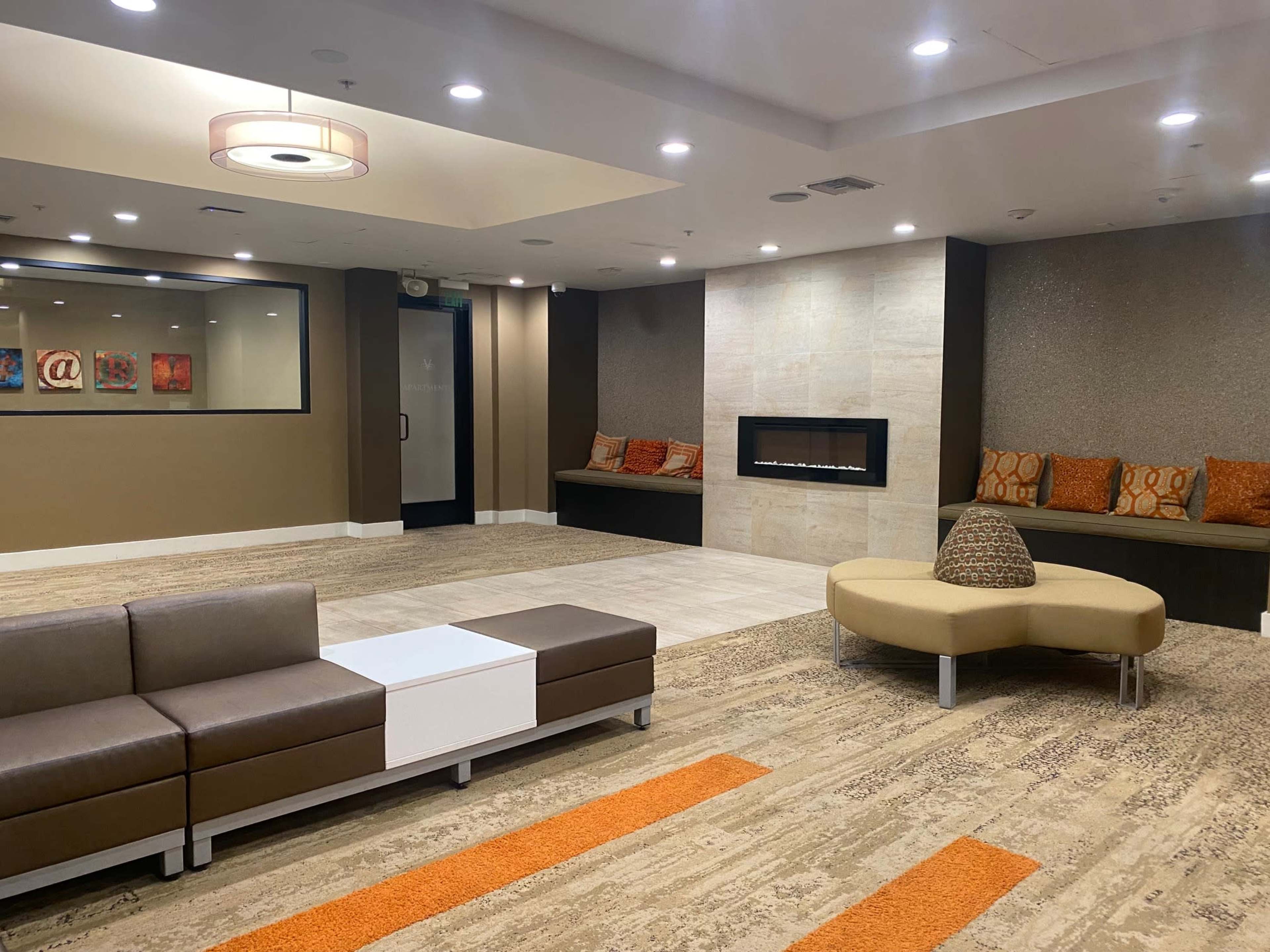 The image shows a modern lounge area with a seating arrangement, a decorative fireplace, and a combination of carpet and tiled flooring.