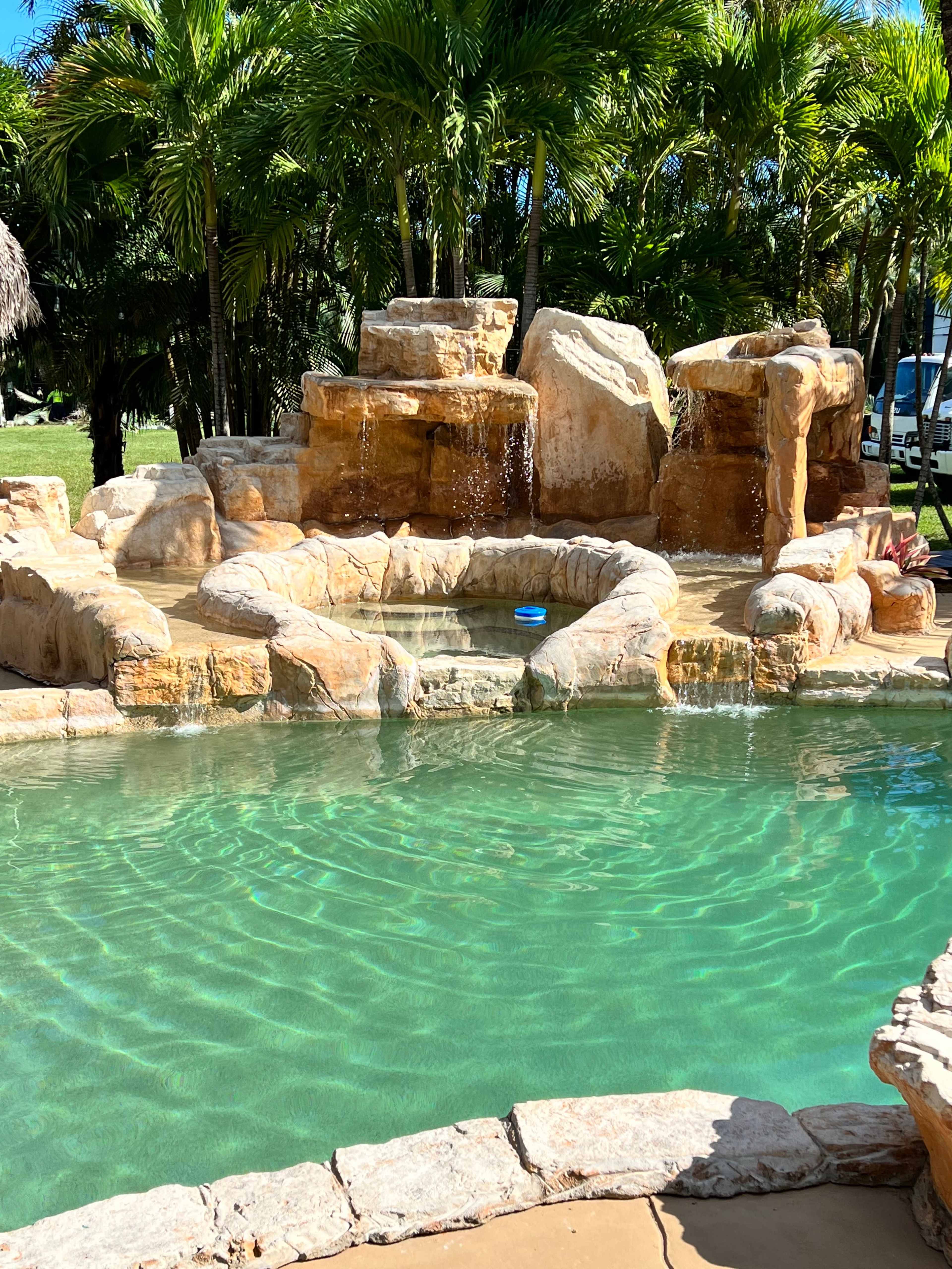 A rocky pool area with waterfalls, surrounded by palm trees and clear blue water.