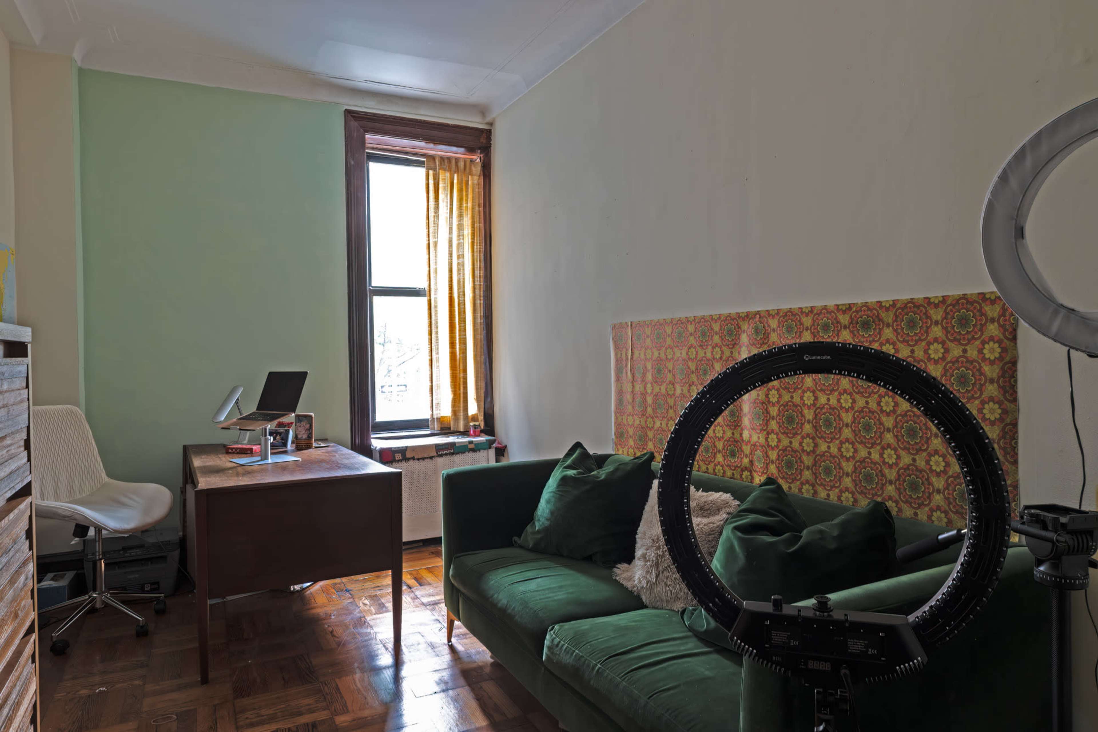 The image shows a small room featuring a green sofa, a wooden desk with a laptop, and a ring light near a window with partially drawn curtains.