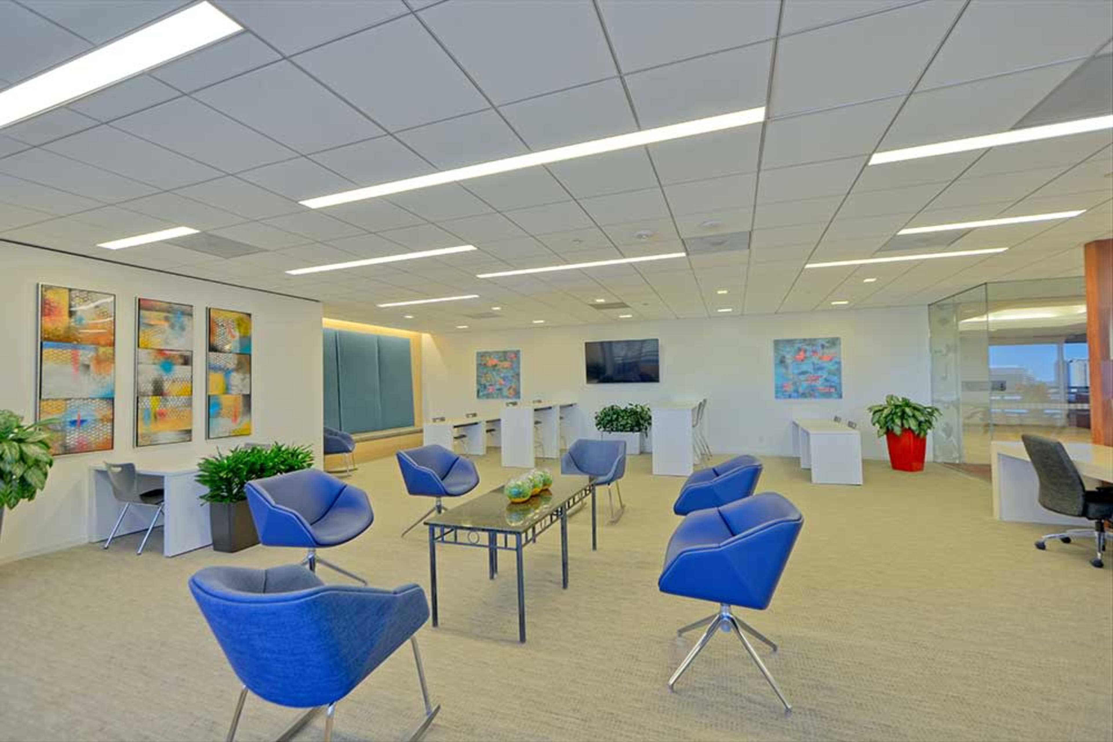 A modern office lobby with blue chairs, colorful artwork on the walls, and several small tables amid plants and a television.