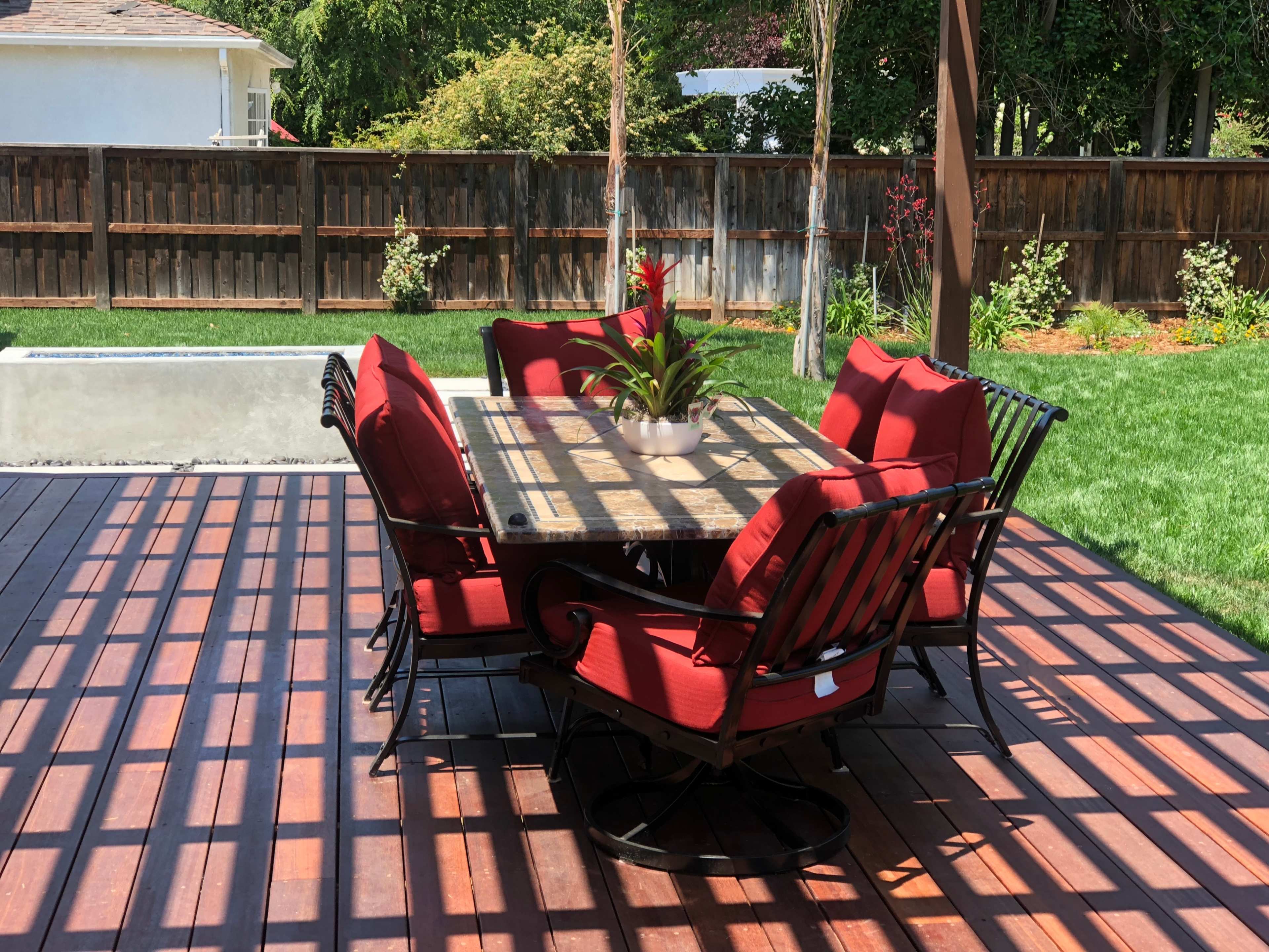 A patio features a table surrounded by chairs with red cushions, set on a wooden deck in a backyard with a green lawn and a wooden fence.