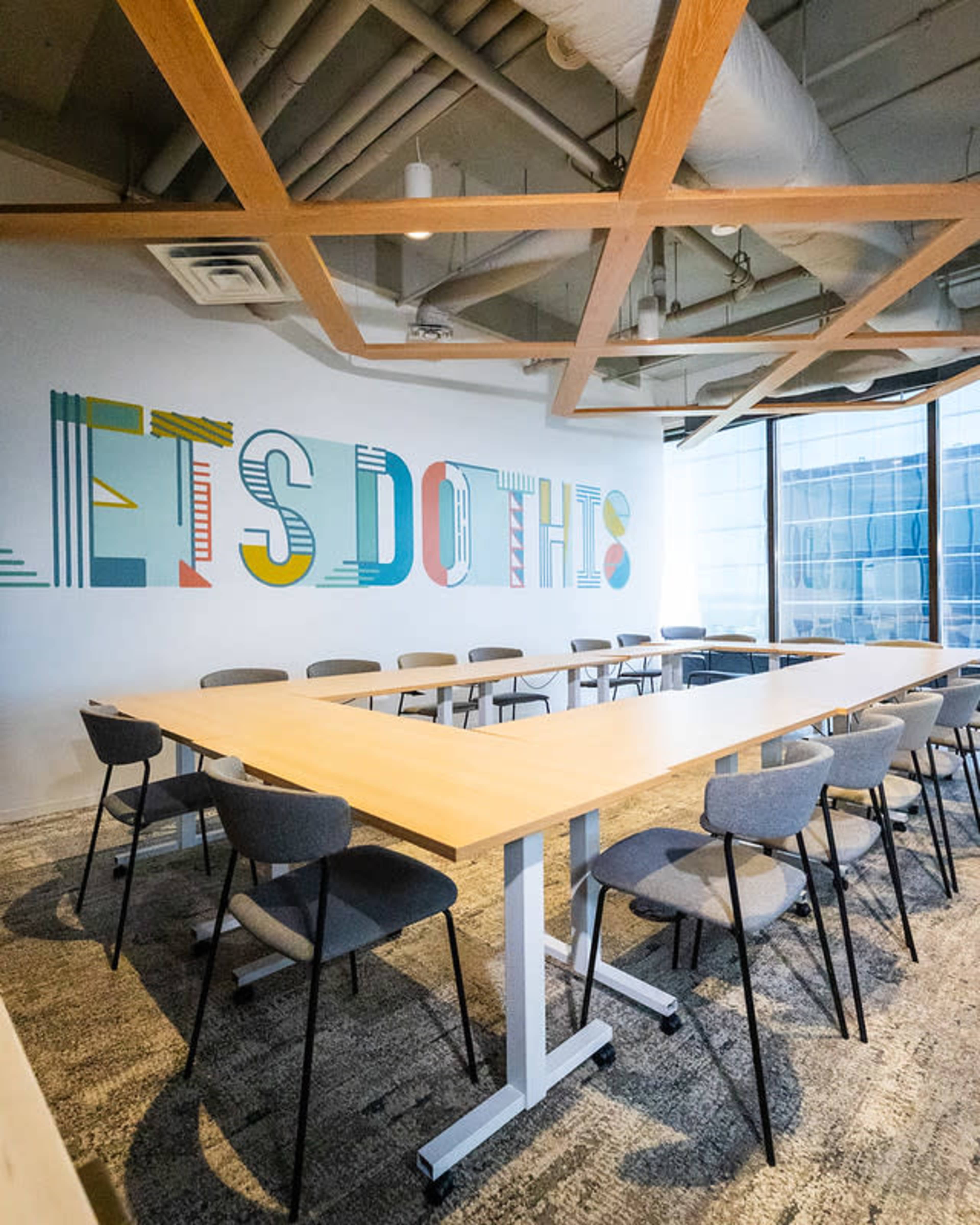 A modern meeting room features a large wooden table surrounded by gray chairs, with a colorful mural on the wall that reads "LET'S DO THIS."