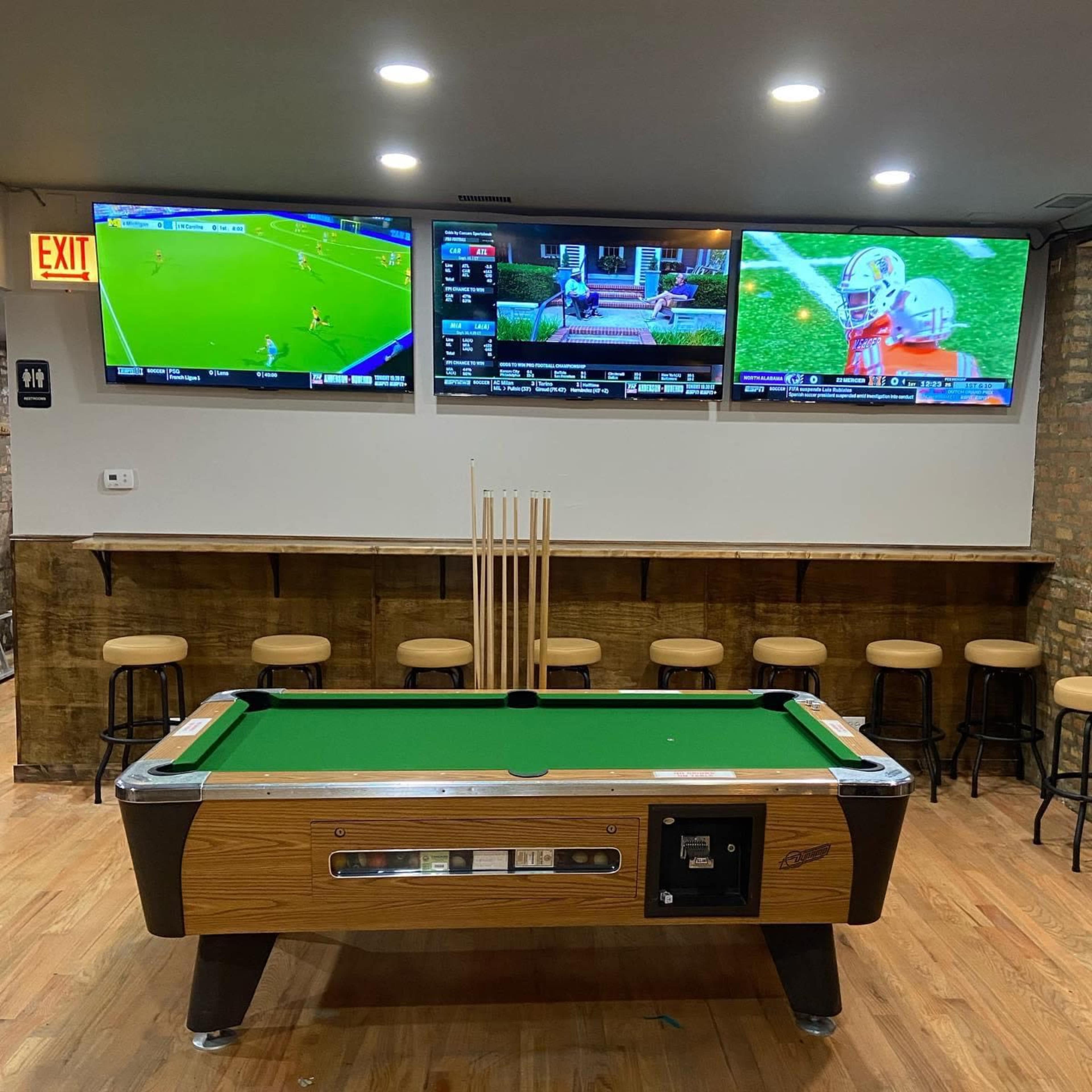 The image shows a bar area with a pool table in the foreground and three television screens displaying sports events on the wall behind it.