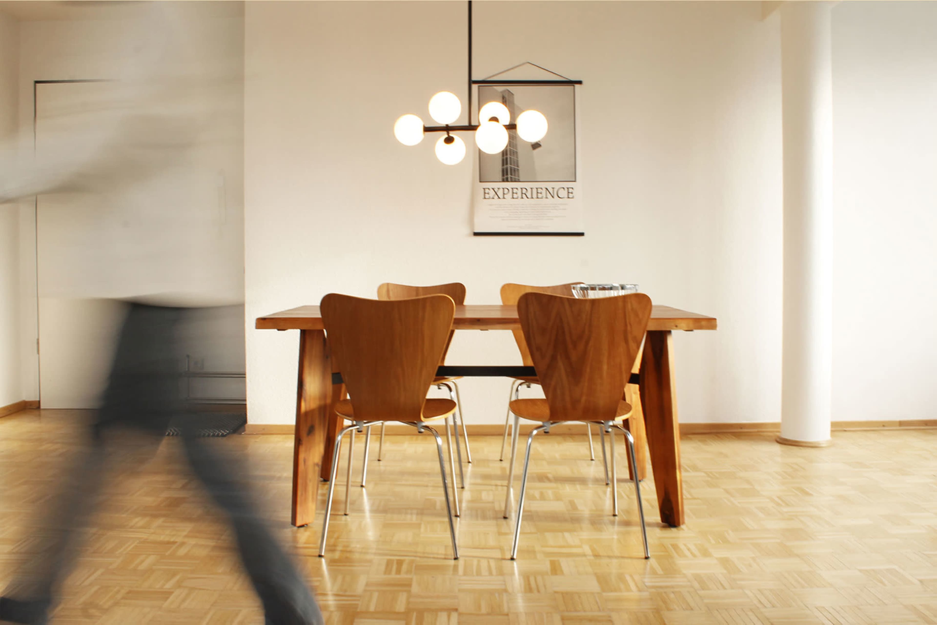A modern dining area features a wooden table with four chairs and a pendant light hanging above, set against a minimalist white wall.