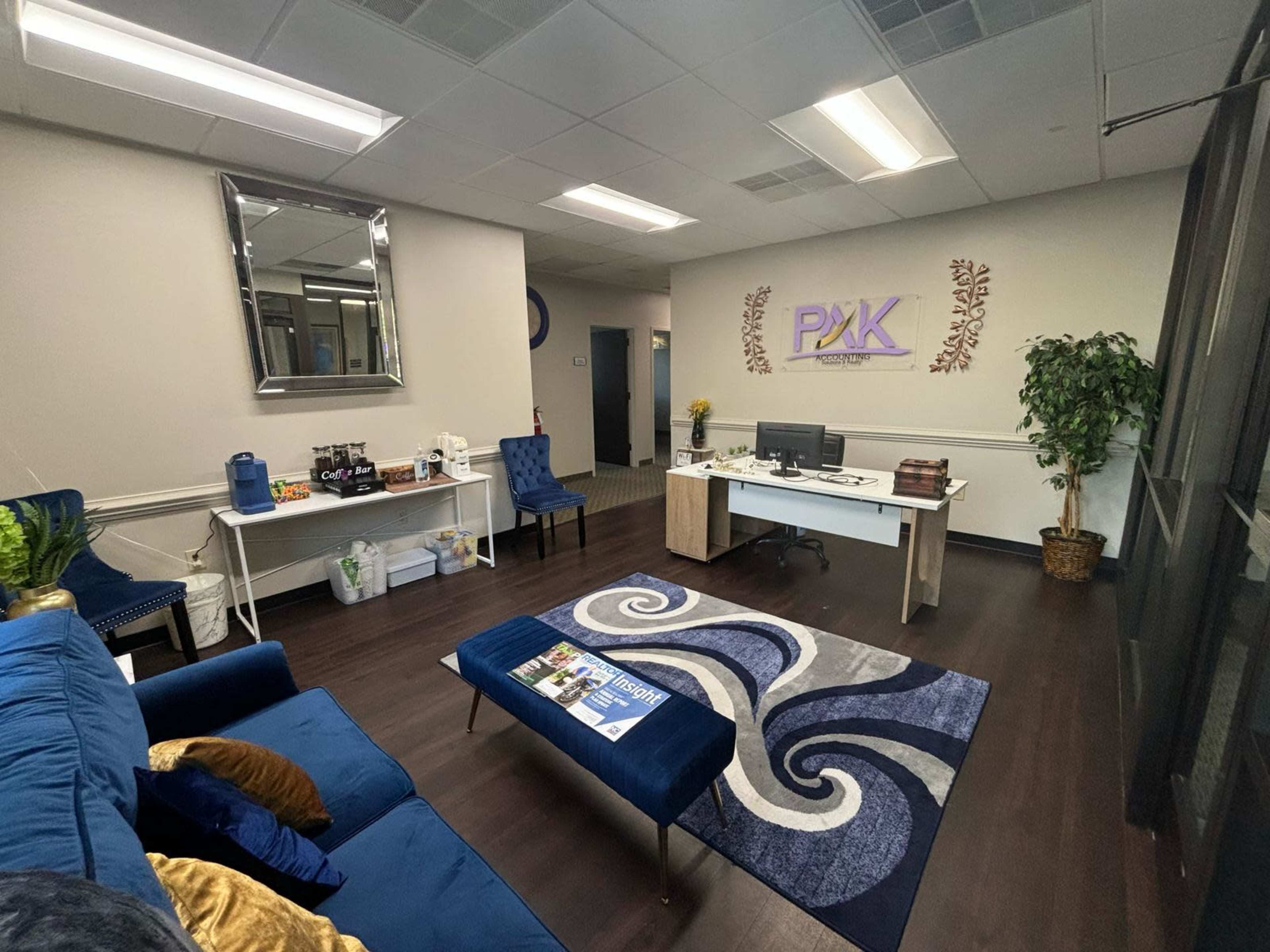 The image shows a modern office space with a reception area featuring a blue couch, a coffee station, a desk, and decorative elements including a rug and wall sign.