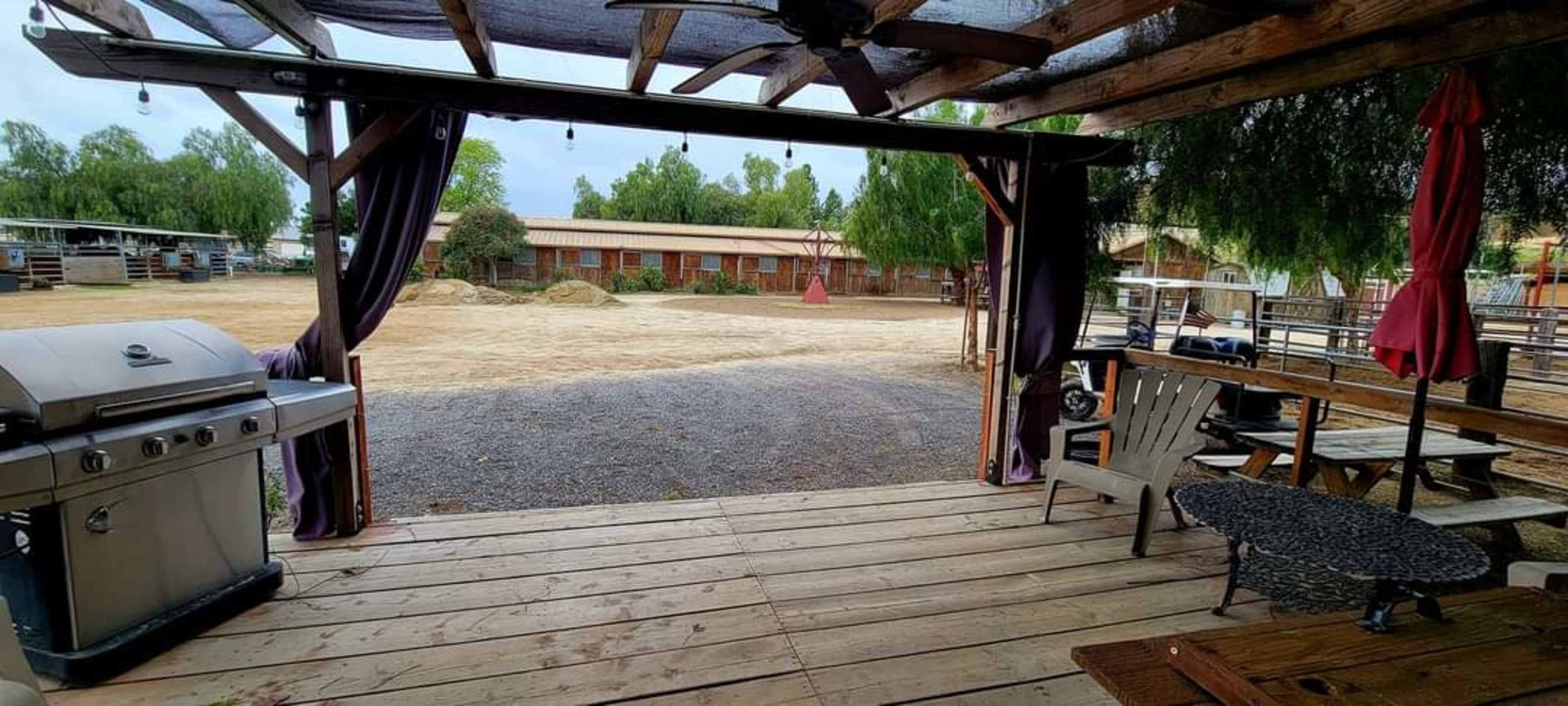 A patio area with a barbecue grill, overlooking a dirt field surrounded by trees and buildings.