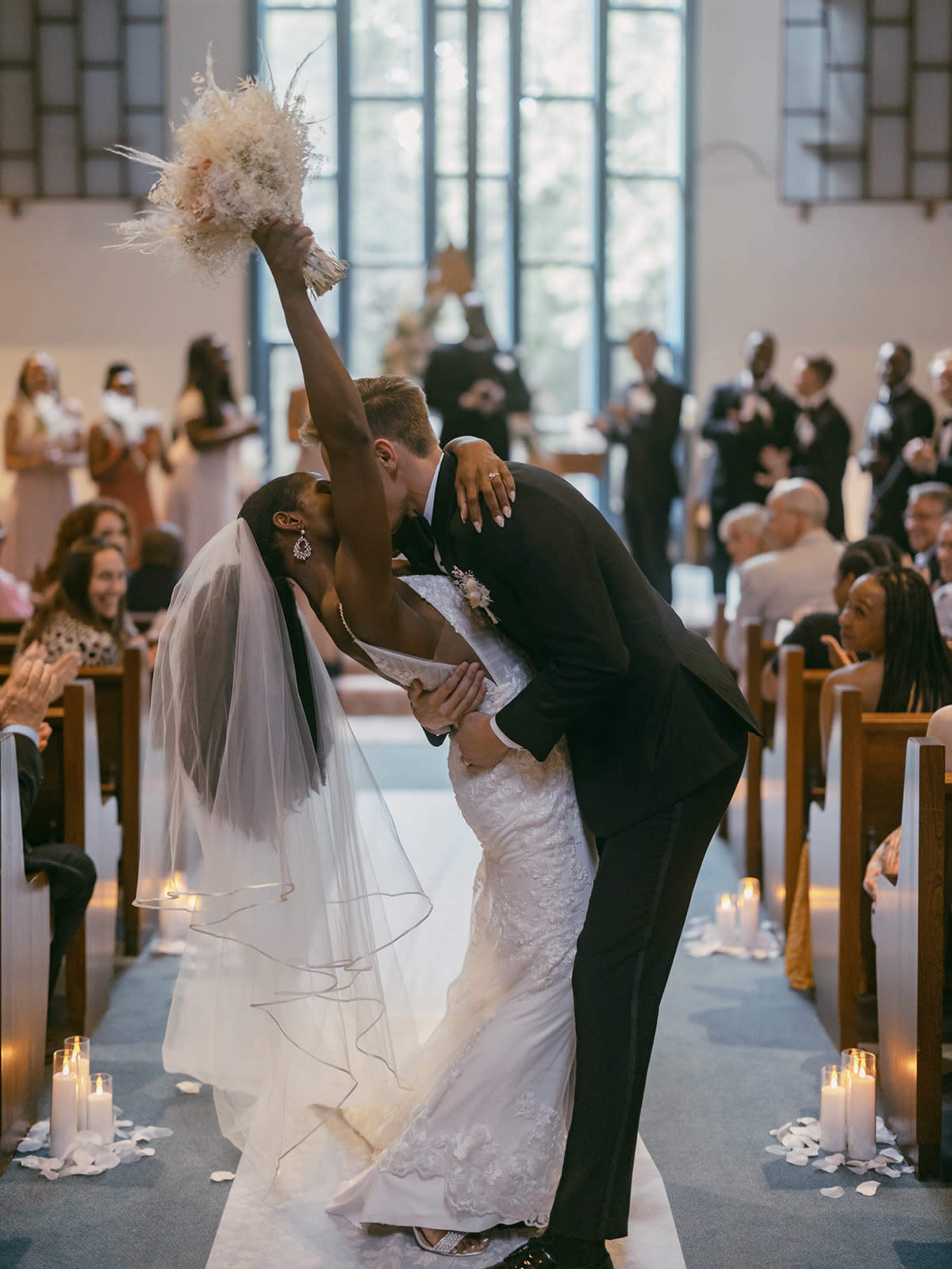 A bride and groom kiss while she raises her bouquet, surrounded by seated guests in a church.