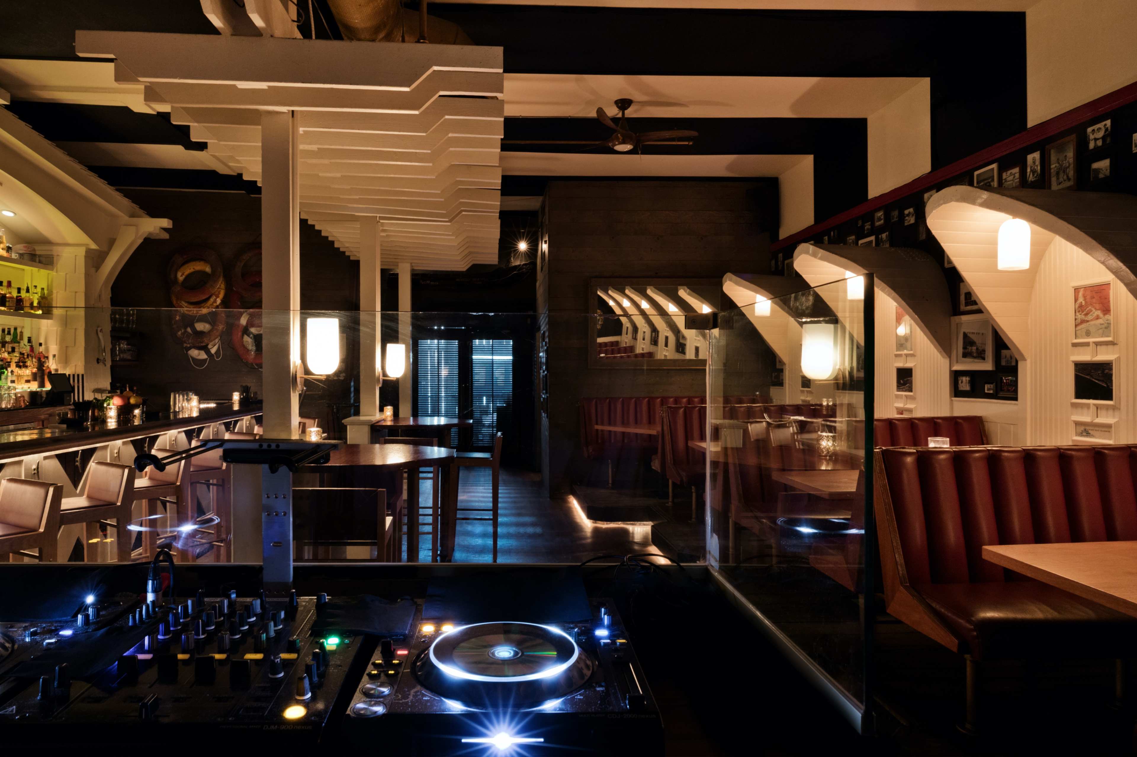 A modern bar and restaurant interior featuring a DJ setup in the foreground with illuminated tables and booths in a dimly lit atmosphere.