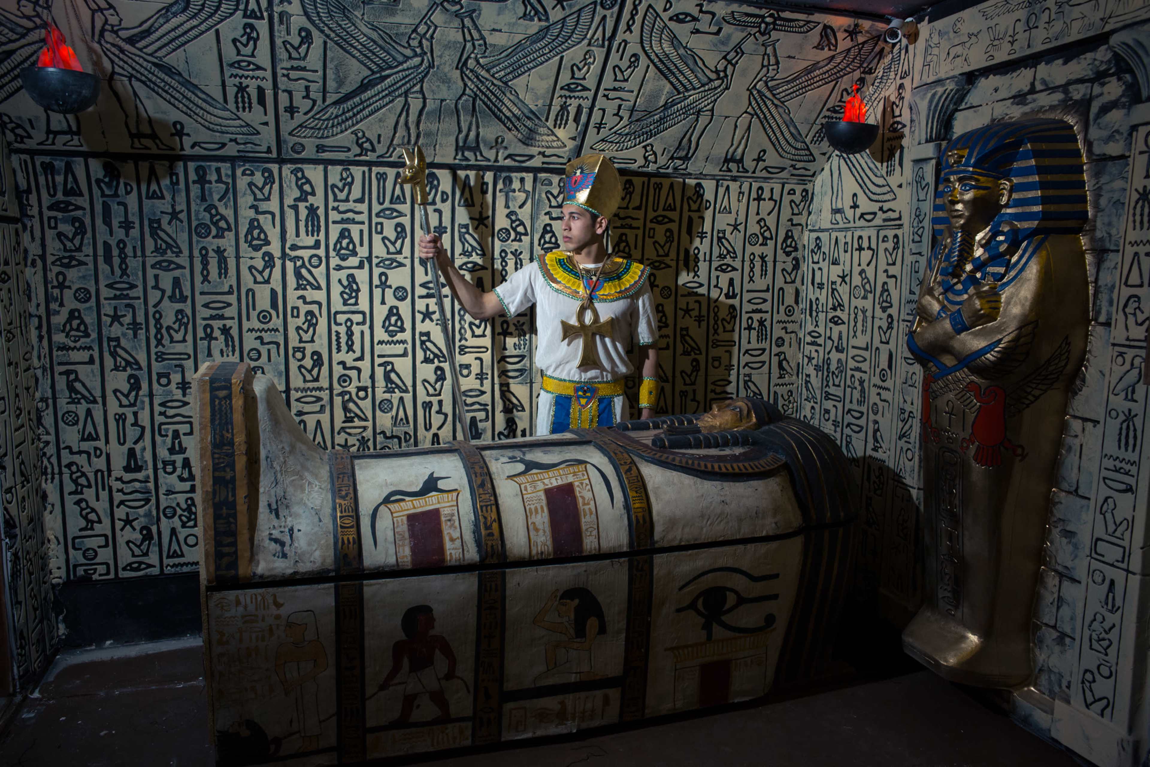 A person in Ancient Egyptian attire stands beside a decorated sarcophagus in a chamber adorned with hieroglyphics and statues.