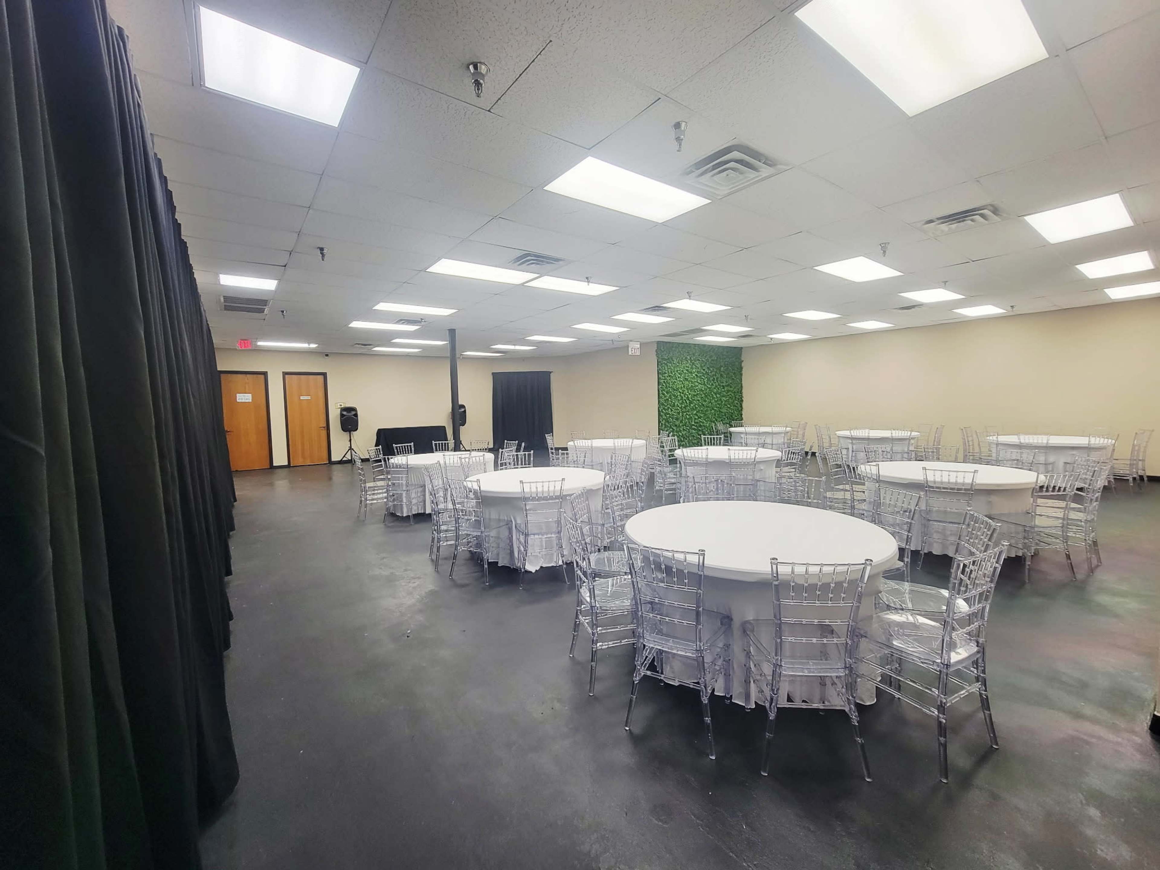 The image shows a spacious event room with multiple round tables adorned with white tablecloths and transparent chiavari chairs, illuminated by bright ceiling lights.