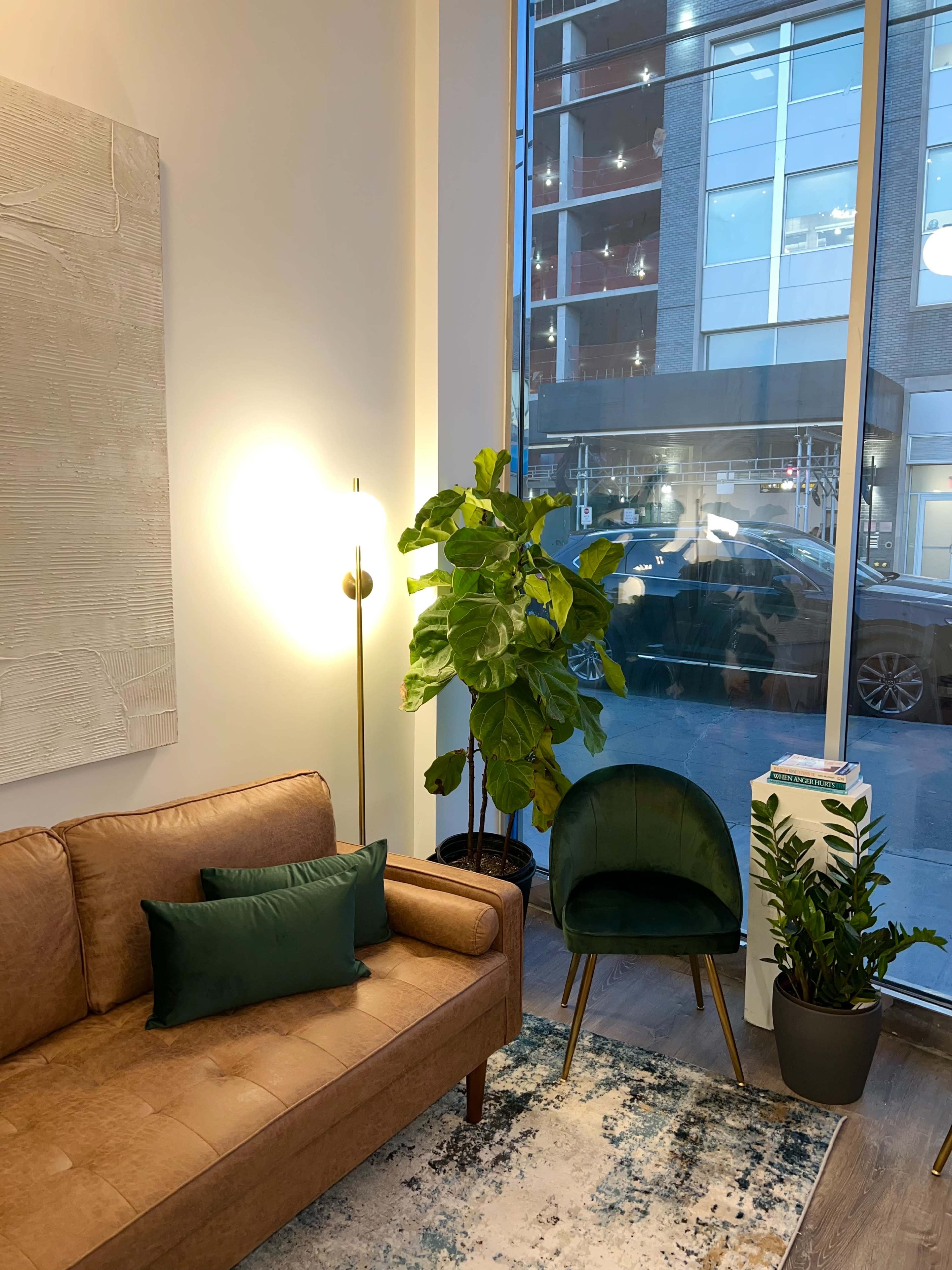 A modern sitting area features a brown sofa, a green chair, indoor plants, and a large window displaying the exterior of a building.