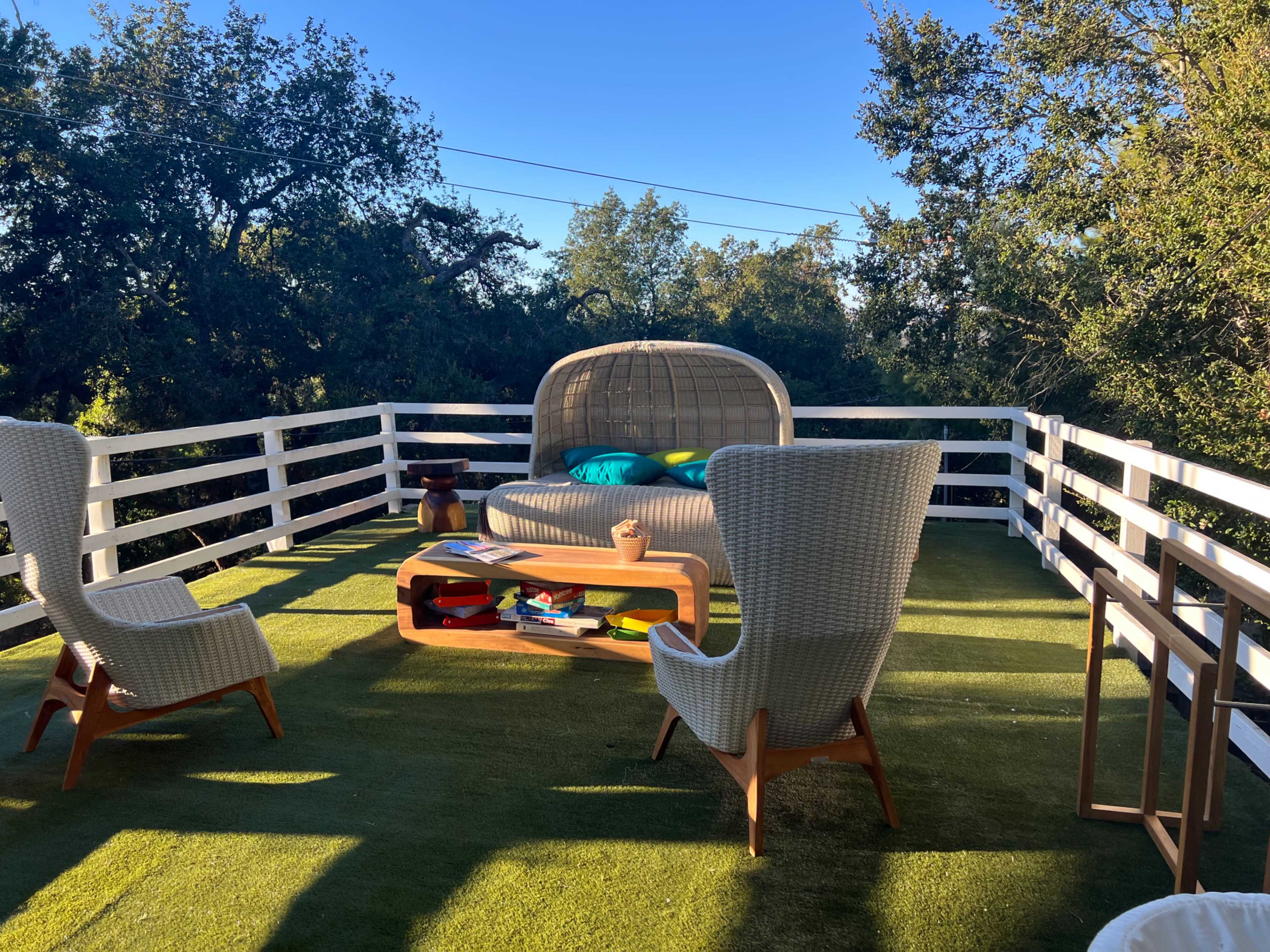 The image shows a cozy outdoor seating area with two armchairs and a central table, surrounded by greenery and white railings.