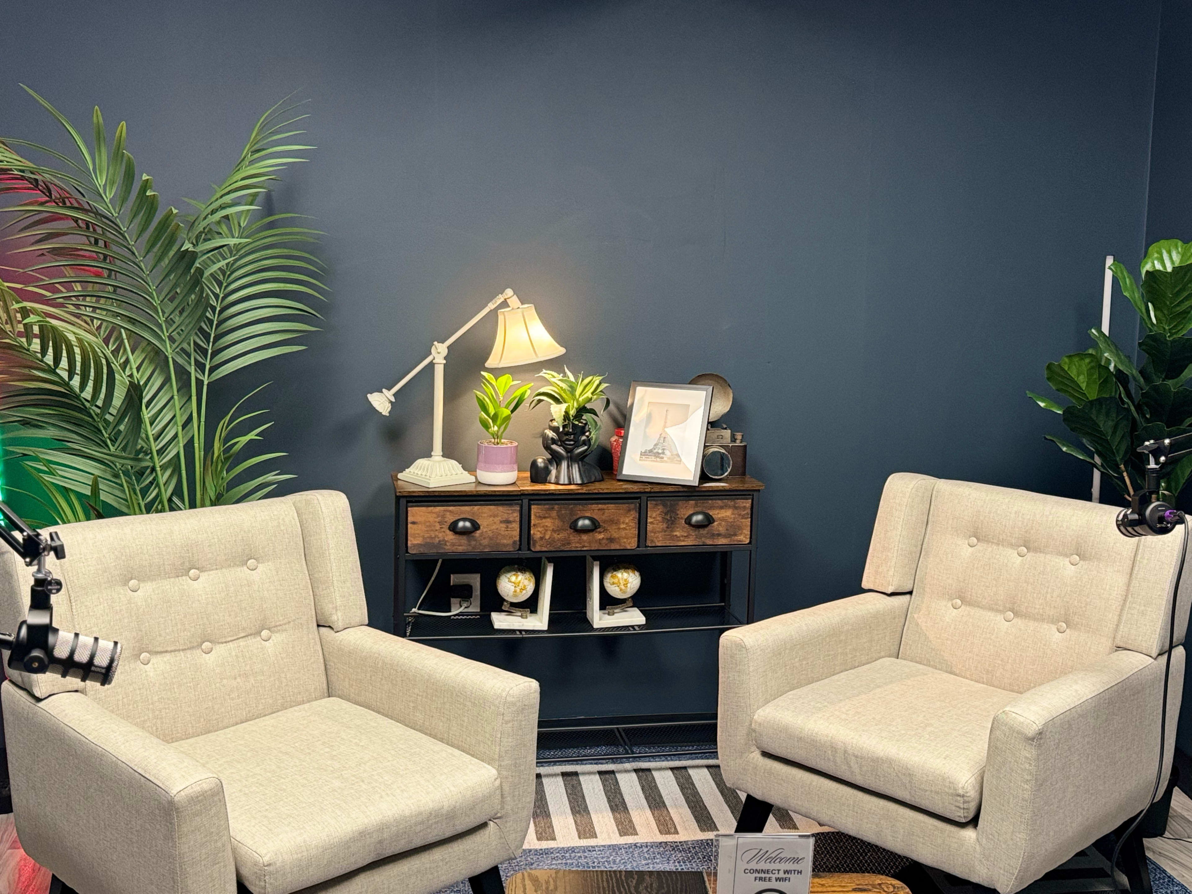 The image shows a cozy setup with two light-colored chairs facing a wooden console table adorned with plants and a lamp, set against a dark wall.