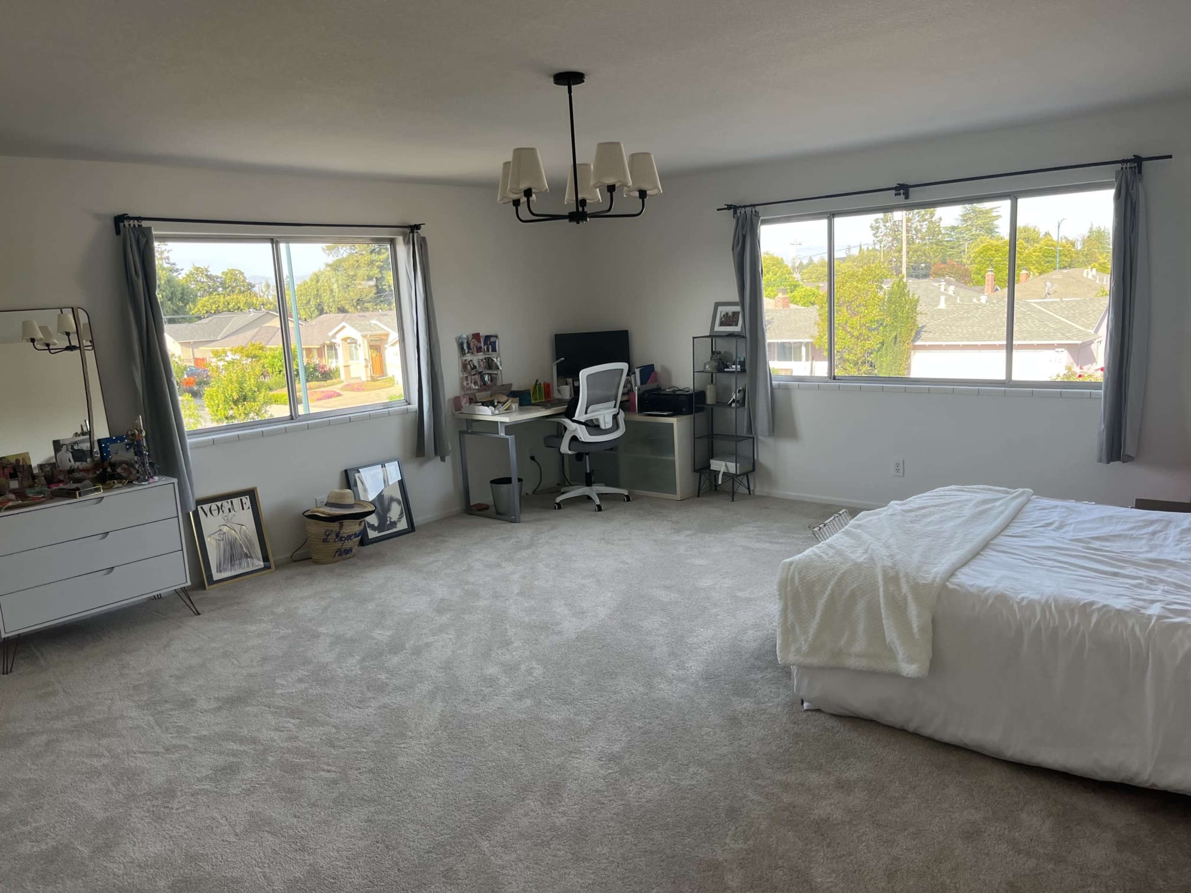 The spacious bedroom features large windows, a desk with a computer, and a neatly made bed.