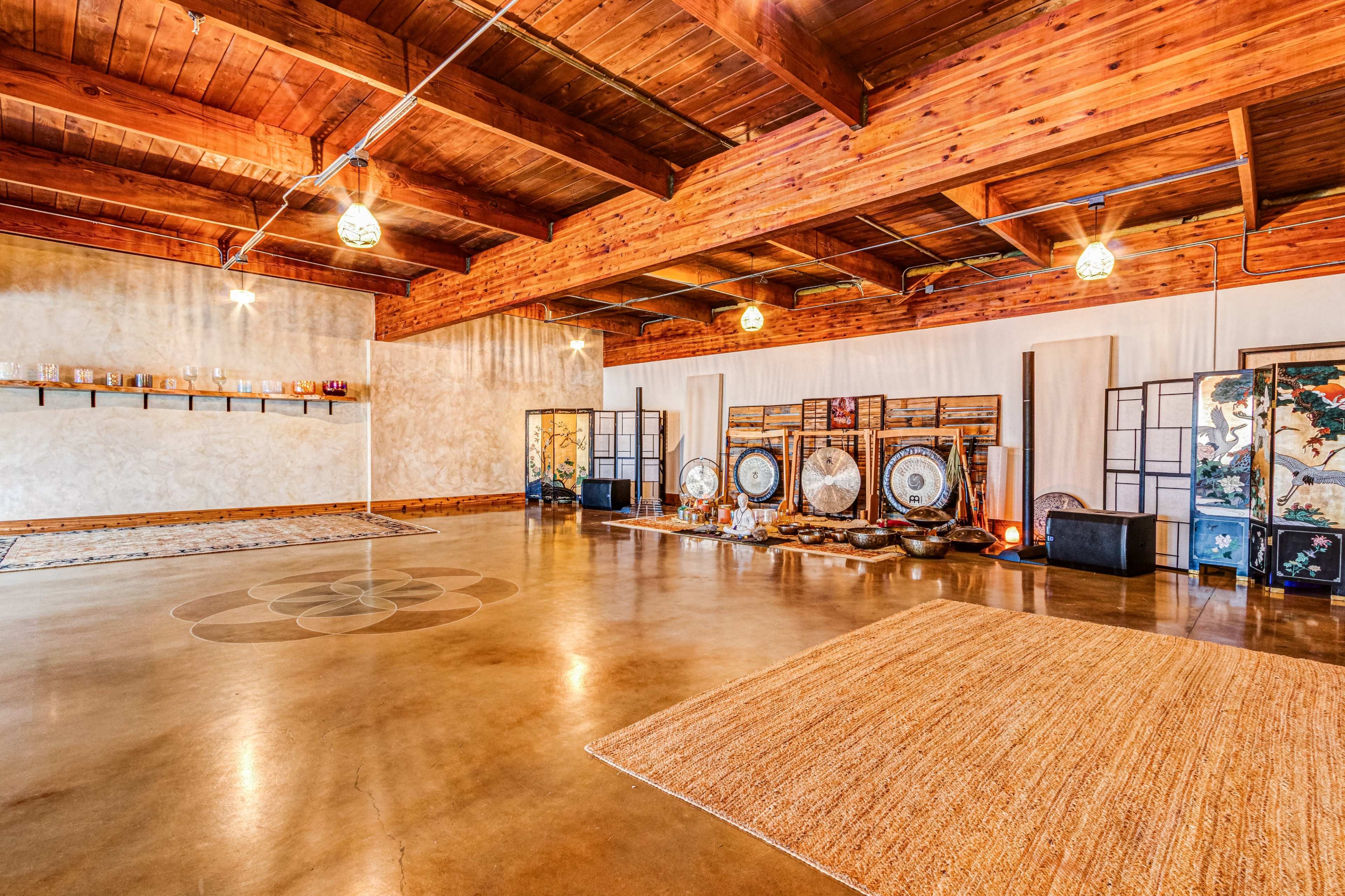 The image shows a spacious meditation or wellness studio with wooden beams, polished concrete flooring, and various instruments arranged against the walls.