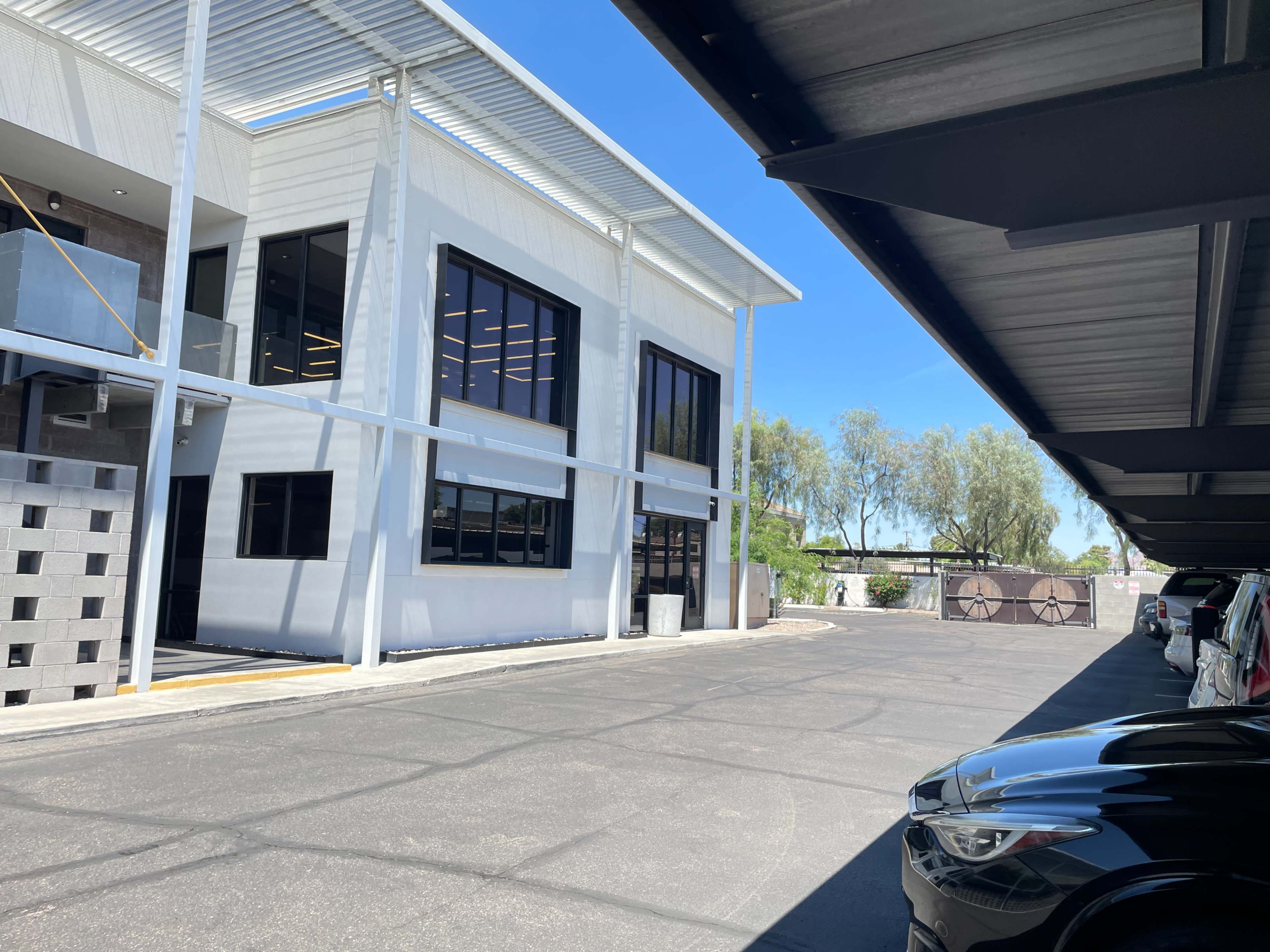 The image shows a modern building with large windows, located adjacent to a paved parking area under a shaded structure.