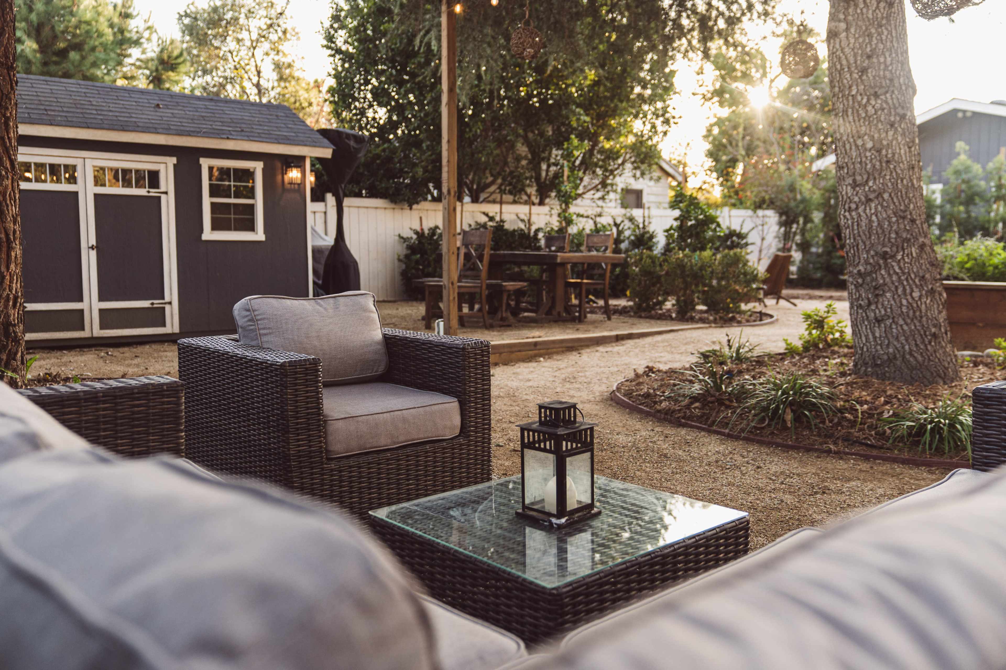 The image shows a cozy backyard seating area with wicker chairs, a glass coffee table, and a gray shed in the background, surrounded by greenery and a dining table.