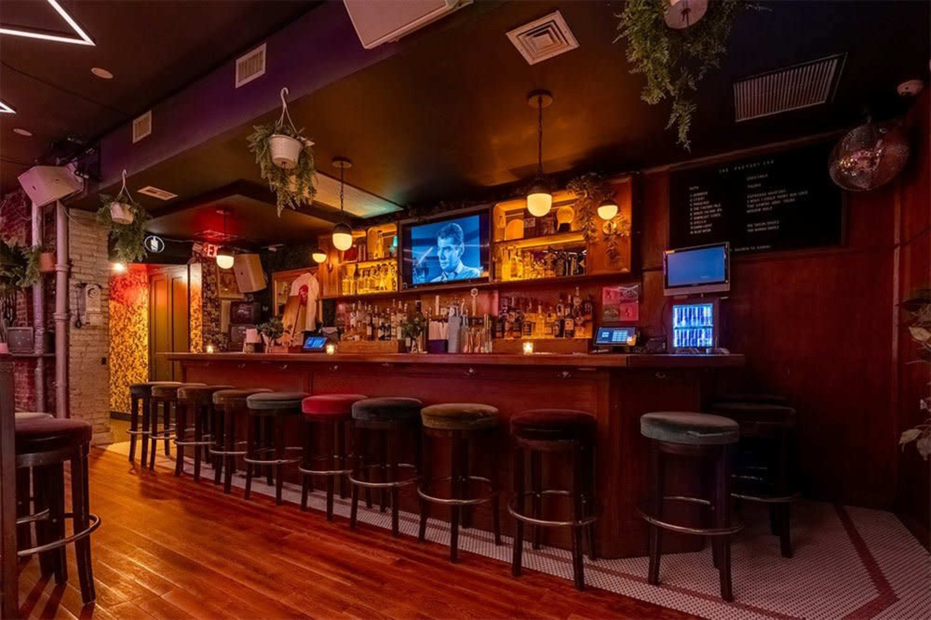 The image shows a dimly lit bar with a long counter, several stools, and a television displaying an image on the wall above.