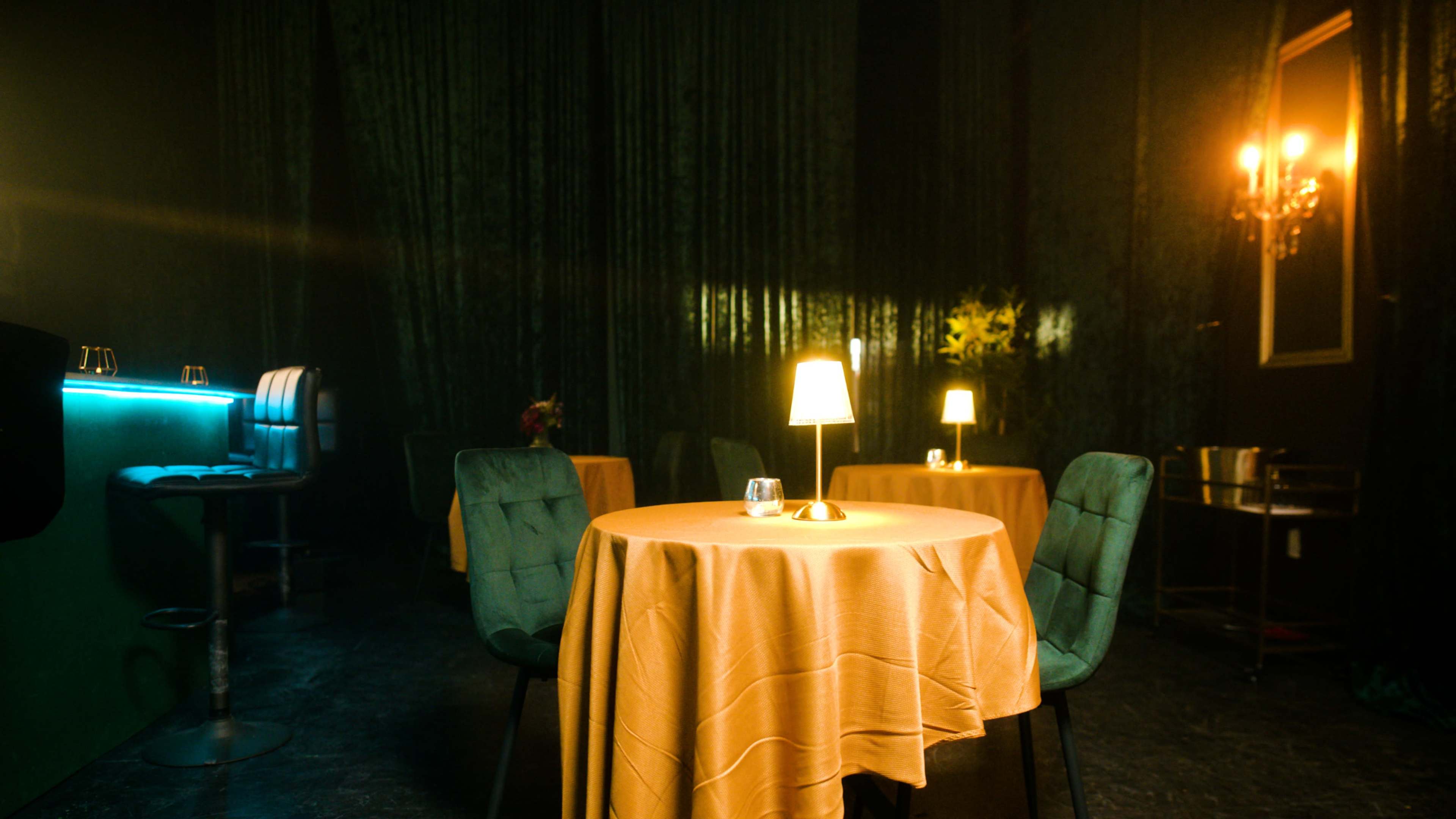 The image shows a dimly lit restaurant scene with round tables covered in yellow tablecloths, each topped with a small lamp and a glass, surrounded by dark velvet curtains.
