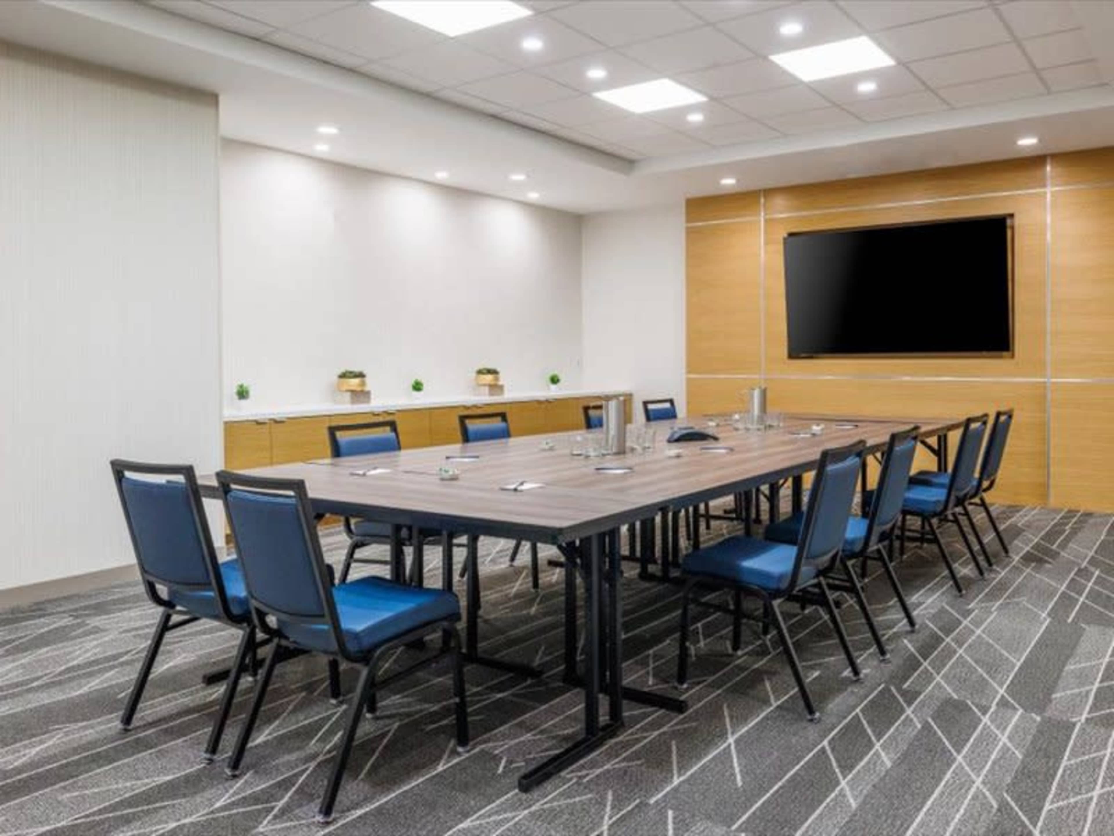 A modern conference room features a large table surrounded by chairs and a wall-mounted screen.